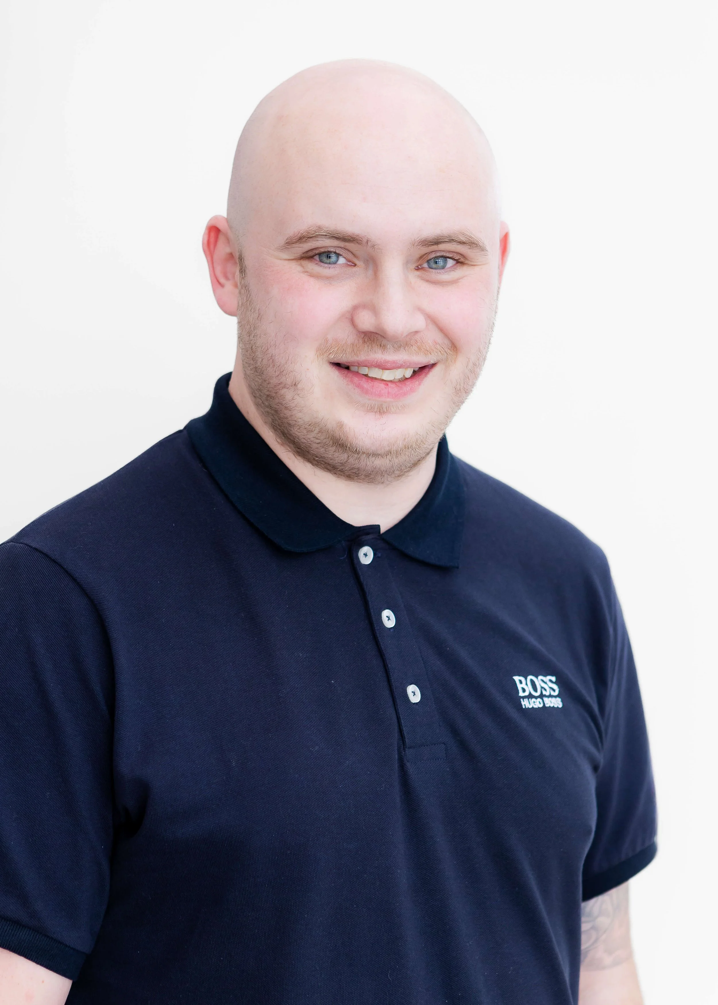 Portrait of a young man with a shaved head and light beard, wearing a dark blue polo shirt with BOSS Hugo Boss logo, against a plain white background.