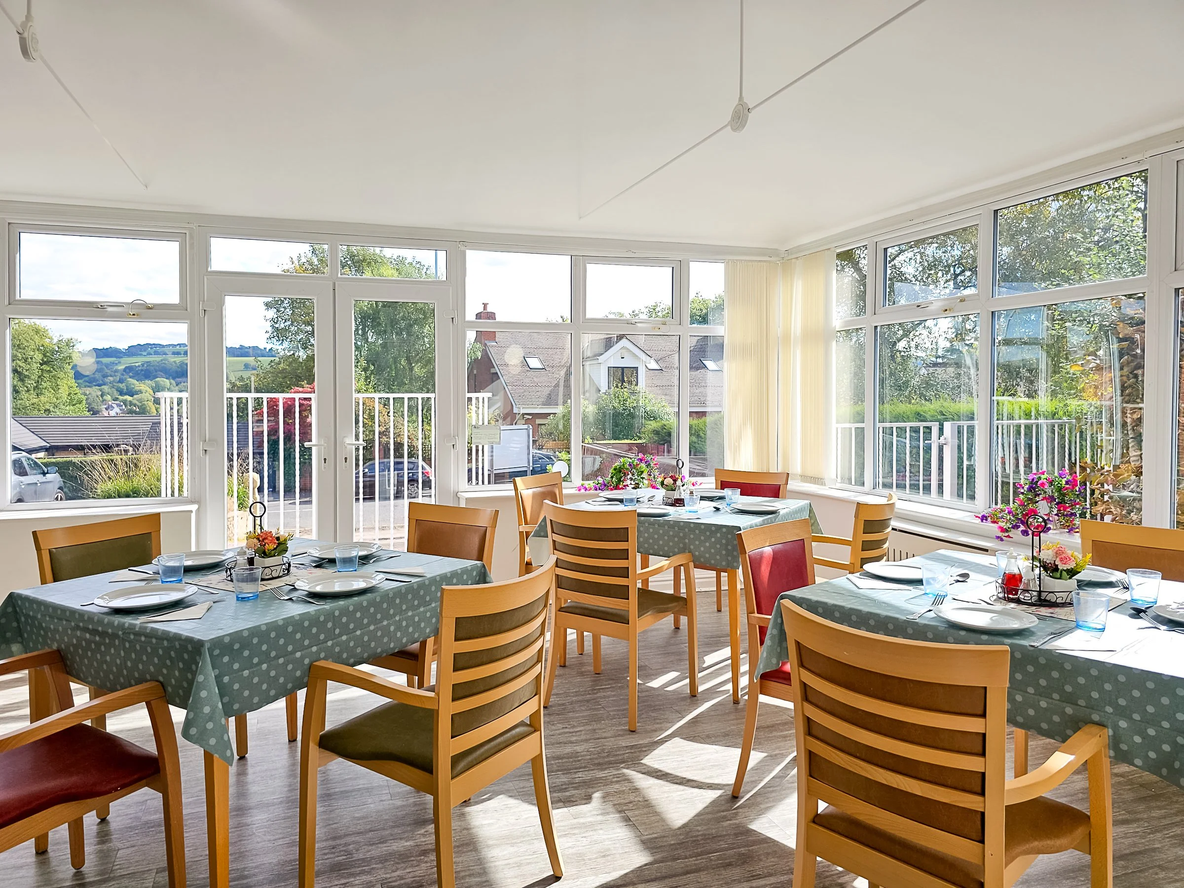 A bright dining area with multiple tables covered in green polka dot tablecloths, set with plates, glasses, and silverware. Large windows let in ample sunlight and offer views of a suburban neighborhood and greenery outside.