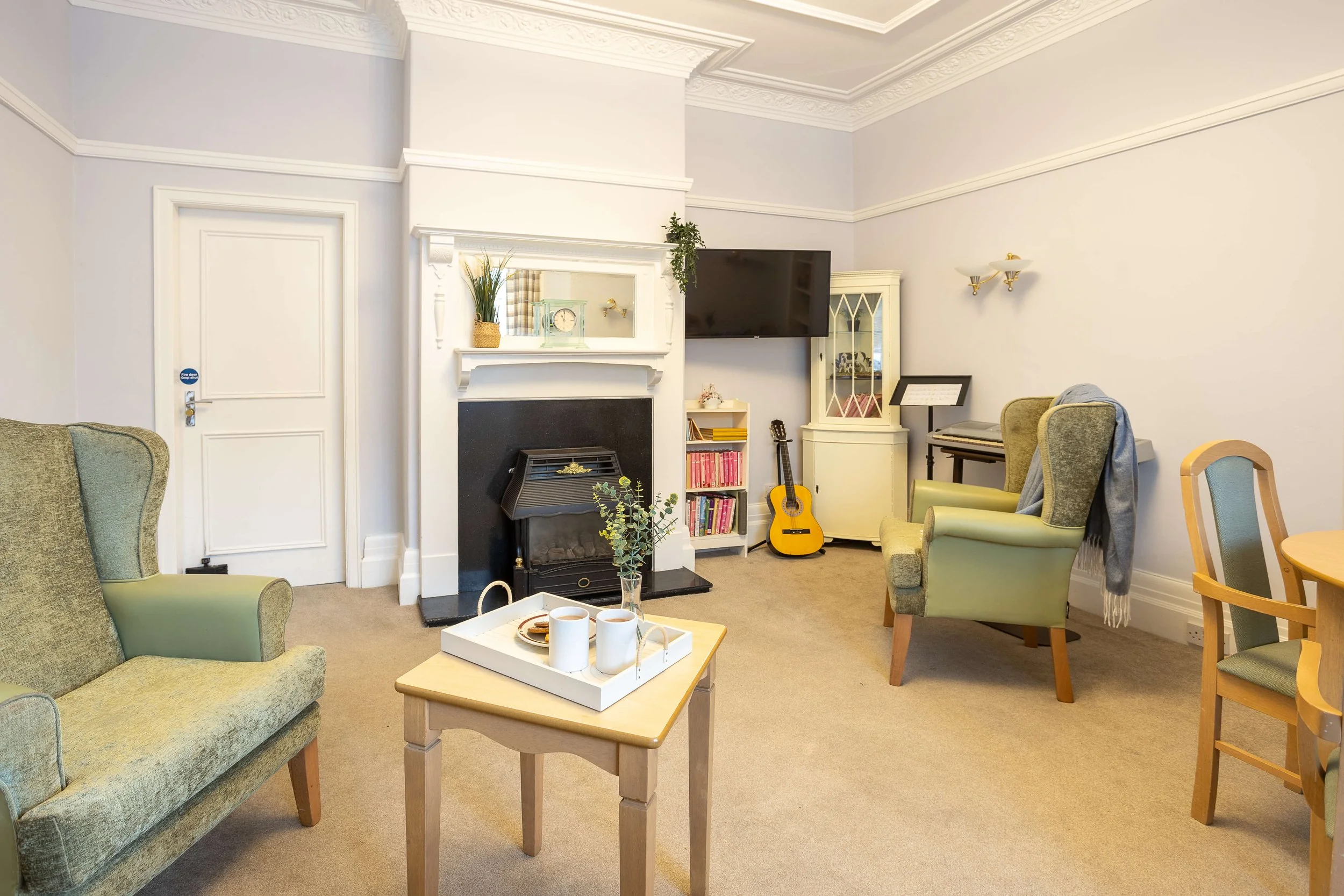 Living room with green and beige armchairs, a small coffee table with a tray of coffee mugs, a fireplace, a mounted TV, a guitar, bookshelf, and piano, with white walls and carpeted floor.