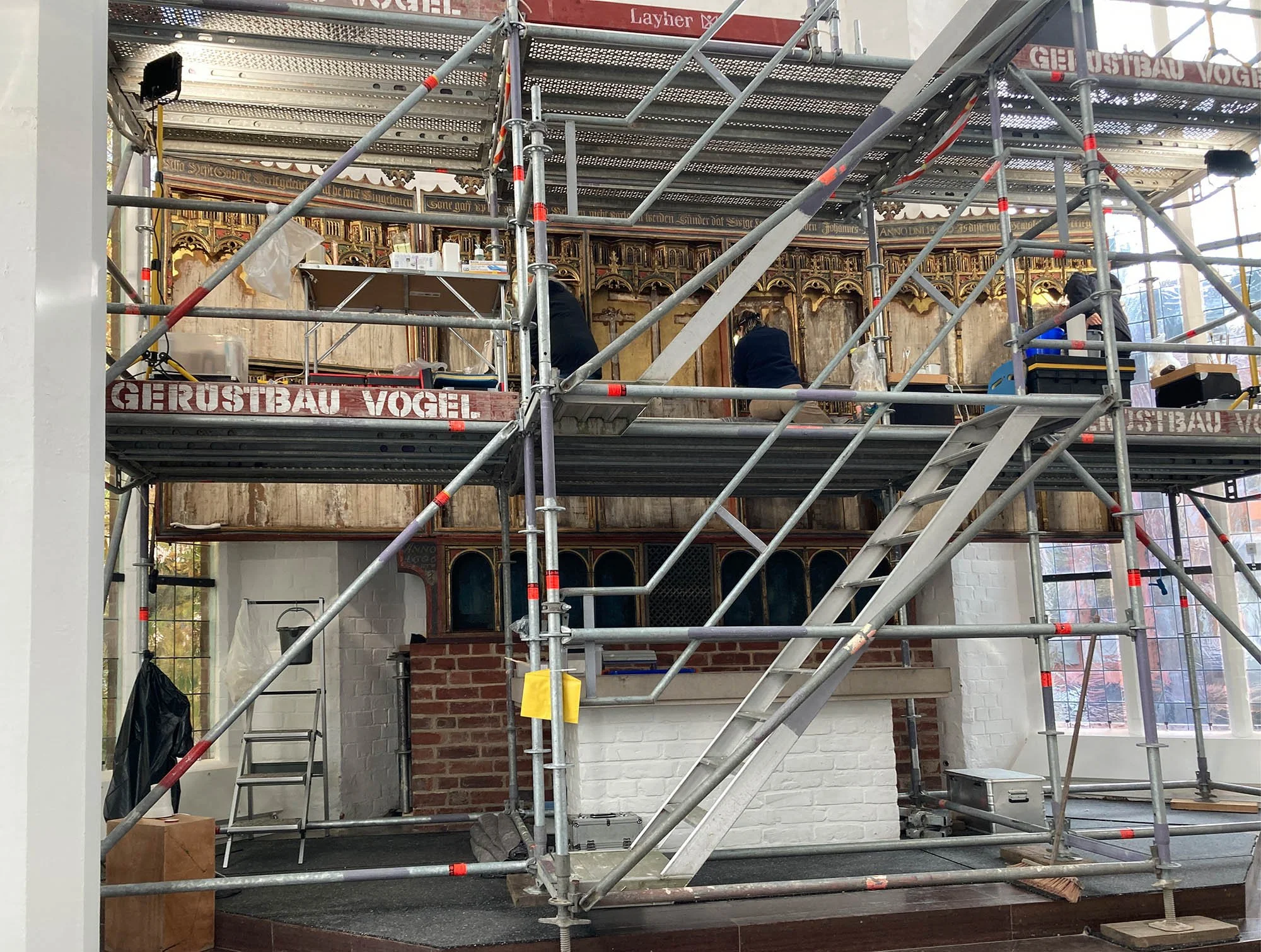 Innenaufnahme einer Kirche oder Kapelle, die derzeit restauriert wird. Es stehen Gerüste, an denen Arbeiter arbeiten, mit Baumaterialien und Werkzeugen. Die Wand im Hintergrund ist kunstvoll mit geschnitzten Holzdetails und einem Altarbereich gestaltet.