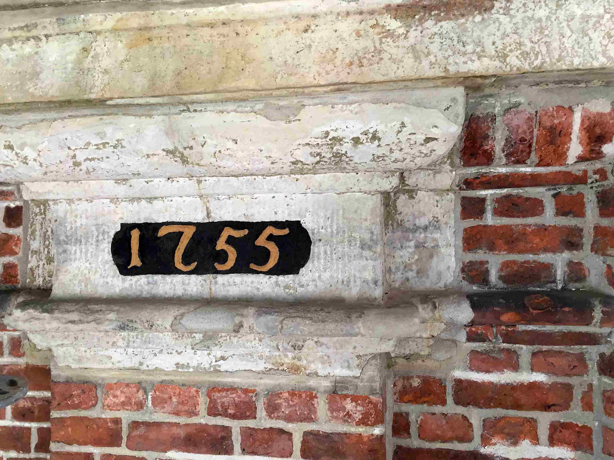 Das Foto zeigt eine alte Stein- und Ziegelmauer mit einer schwarzen Tafel, auf der die Zahlen 1755 in goldener Schrift geschrieben sind.