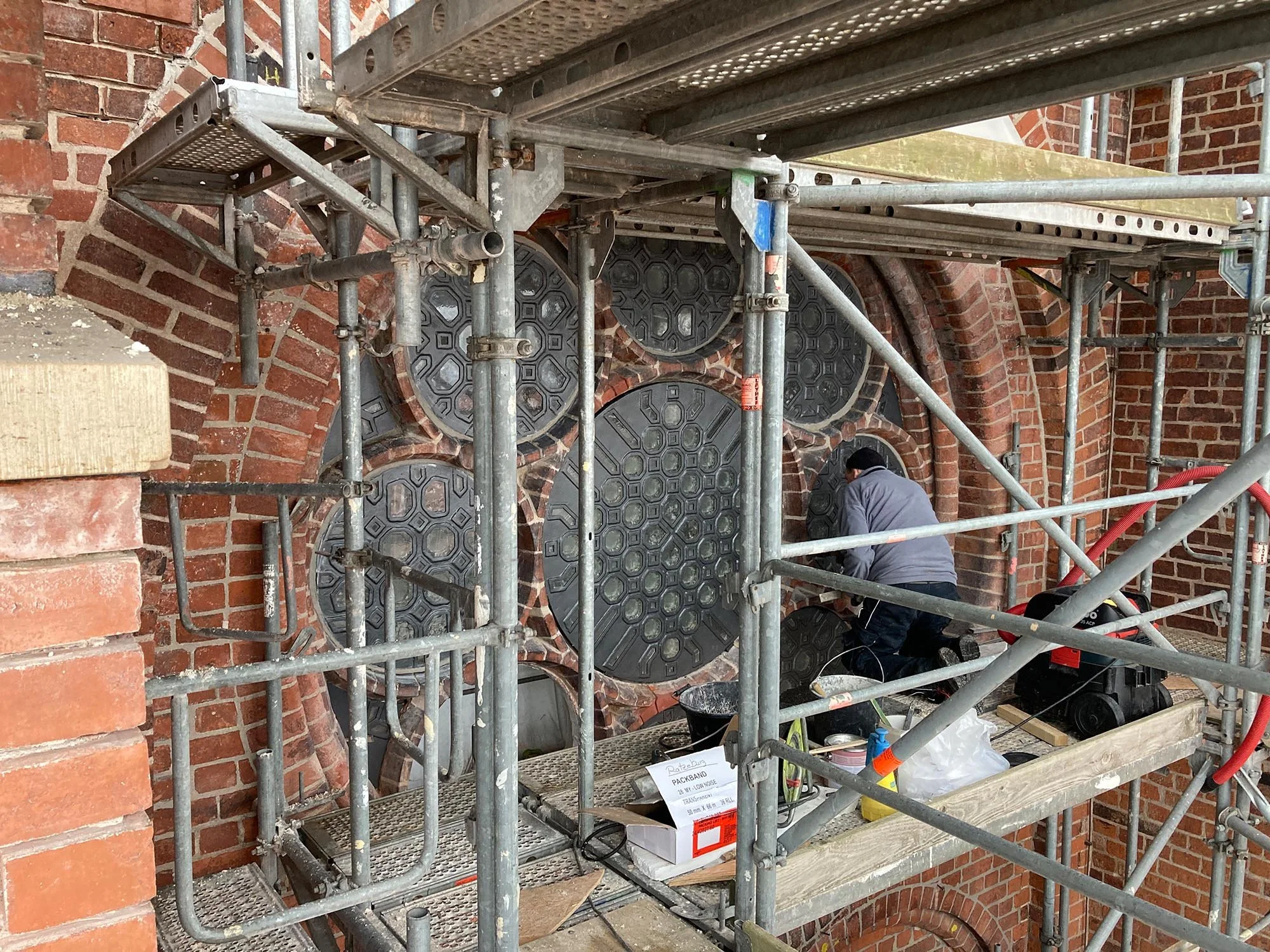 Ein Handwerker arbeitet an einer alten Backsteinmauer mit runden Metallabdeckungen, während er auf einer Gerüstkonstruktion steht.