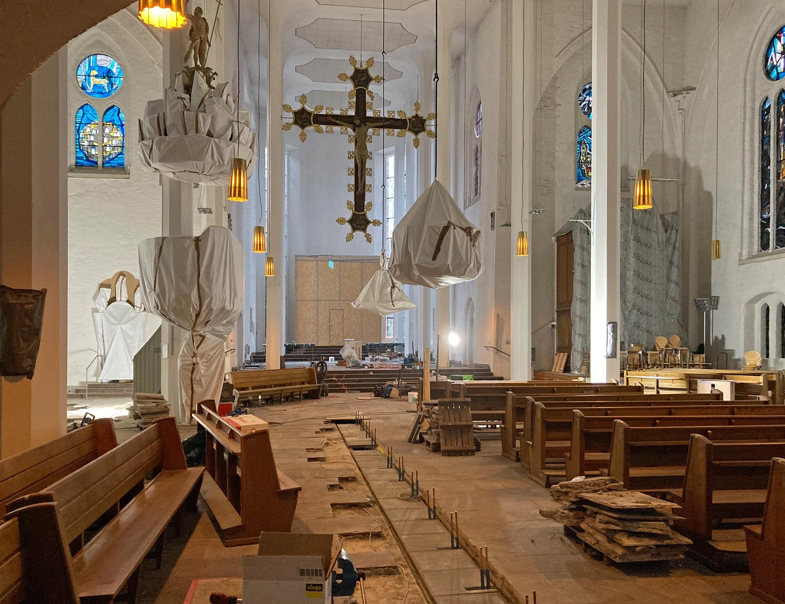 Innenansicht einer Kirche, die renoviert wird, mit Bauarbeiten, Werkzeugen und Baumschutt auf dem Boden, Schutzabdeckungen an den Säulen, und noch nicht vollständig fertiggestellten Bereichen.