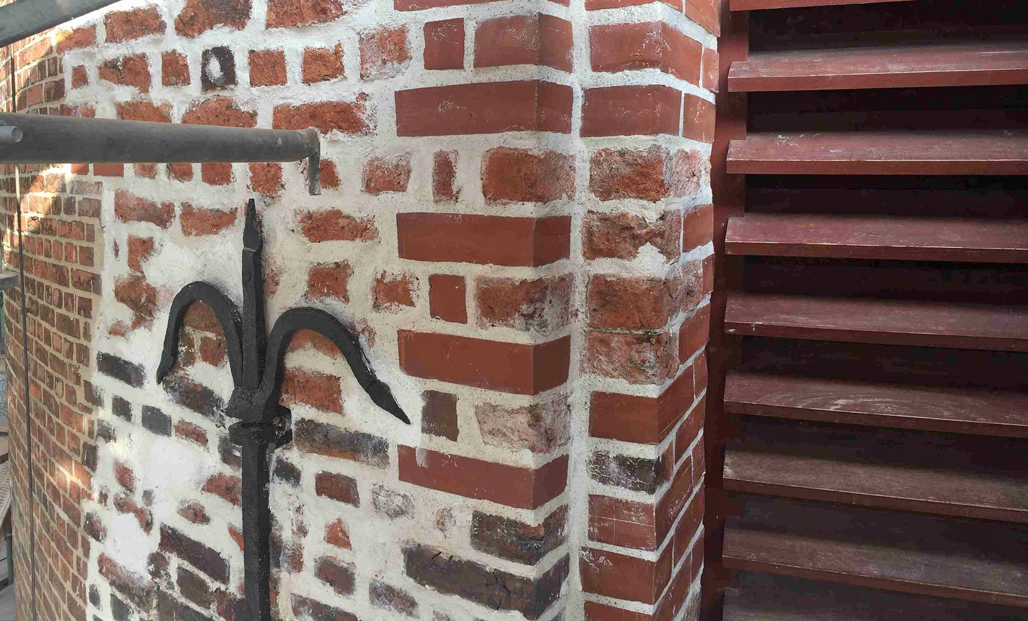 Nahaufnahme eines roten Backsteingebäudes mit Holztreppe auf der rechten Seite und einem schwarzen Metallhaken an der Mauer auf der linken Seite.