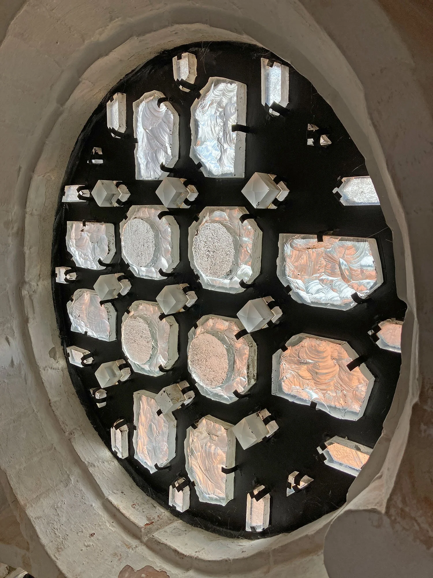 Blick durch ein dekoratives Kirchenfenster mit mehreren kleinen Glasstücken, die durch dunklen Rahmen zusammengehalten werden, mit sichtbaren Bögen und Steinen um das Fenster herum.