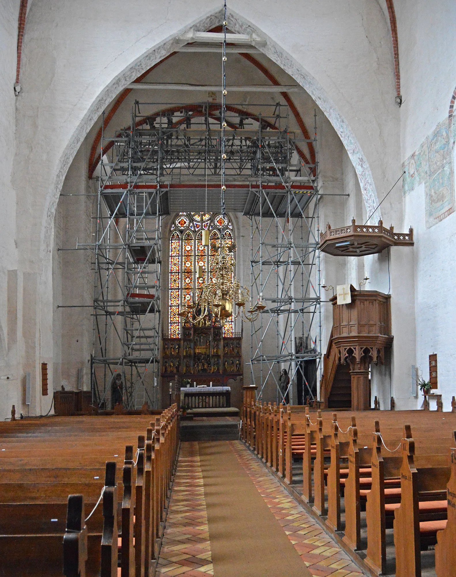 Innenansicht einer Kirche mit Gerüst, vermutlich wegen Renovierungsarbeiten, im vorderen Bereich der Kirche stehen Holzsitzbänke, im Hintergrund ein Altar mit einem Kronleuchter und bunten Glasfenstern.