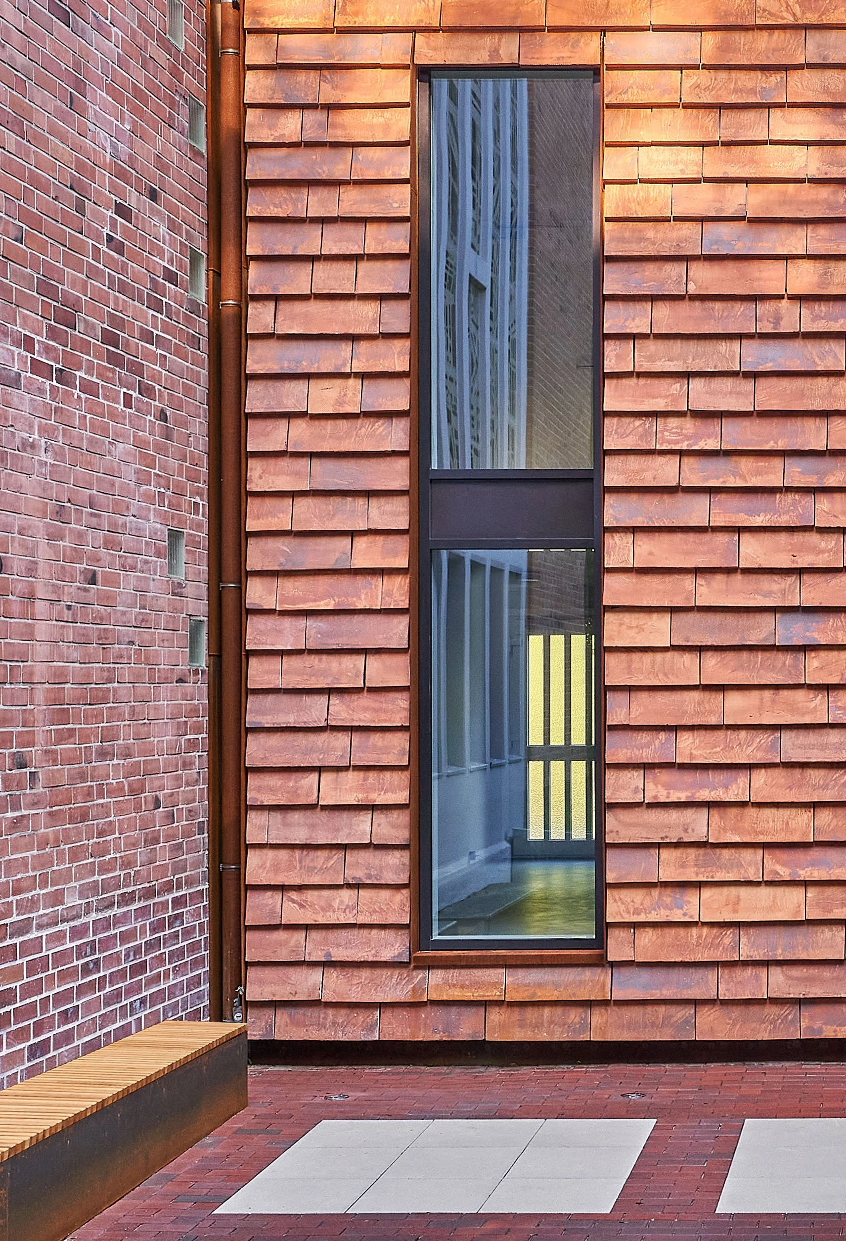 Ansicht einer modernen Gebäudefassade mit roten Ziegeln und vertikaler Fensterfront aus Glas und Metall. Im Vordergrund ein gepflasterter Gehweg.