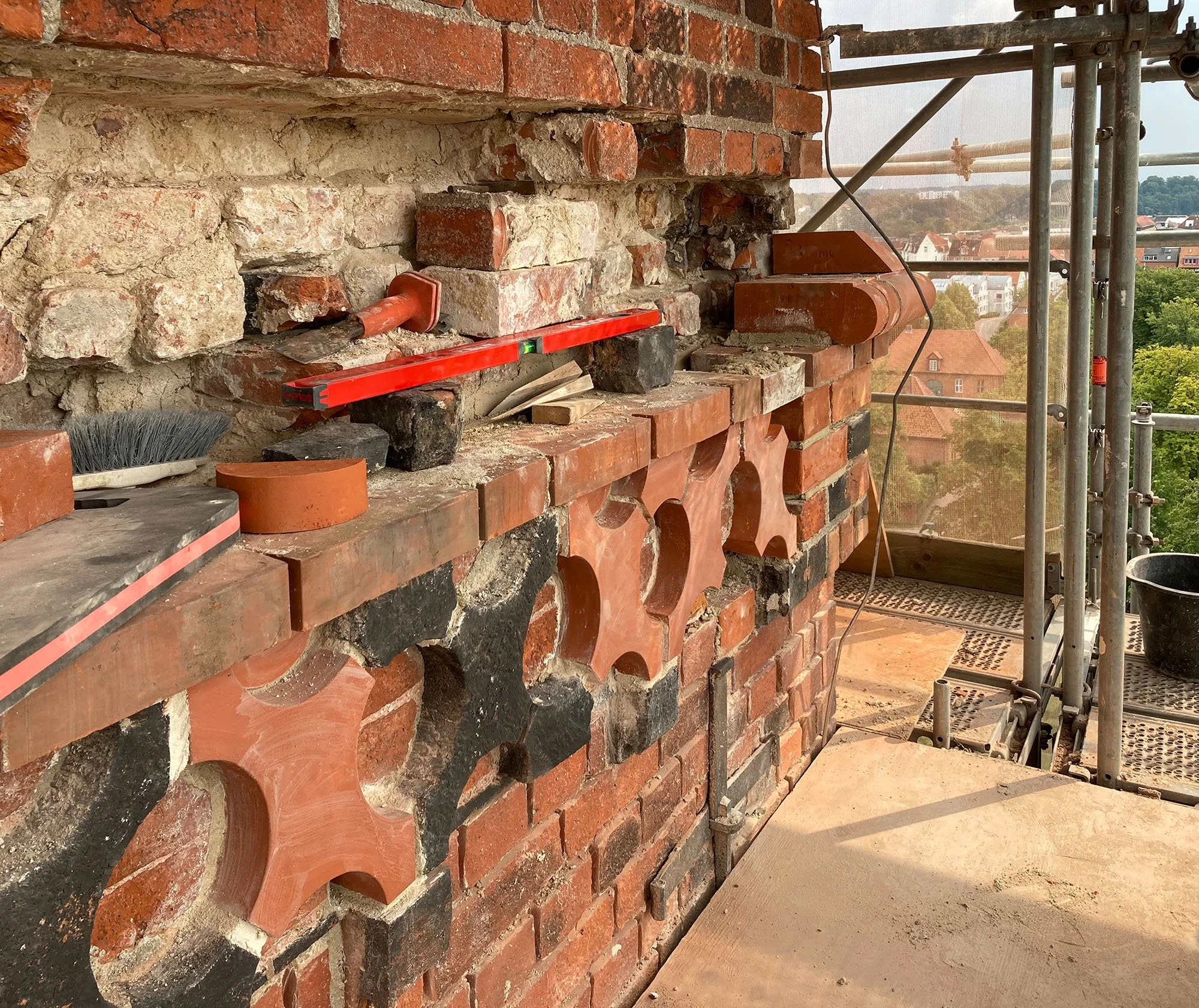 Baustelle mit roten Ziegeln und Baustellengerüst, Aussicht auf eine Stadt mit Häusern und Bäumen im Hintergrund.