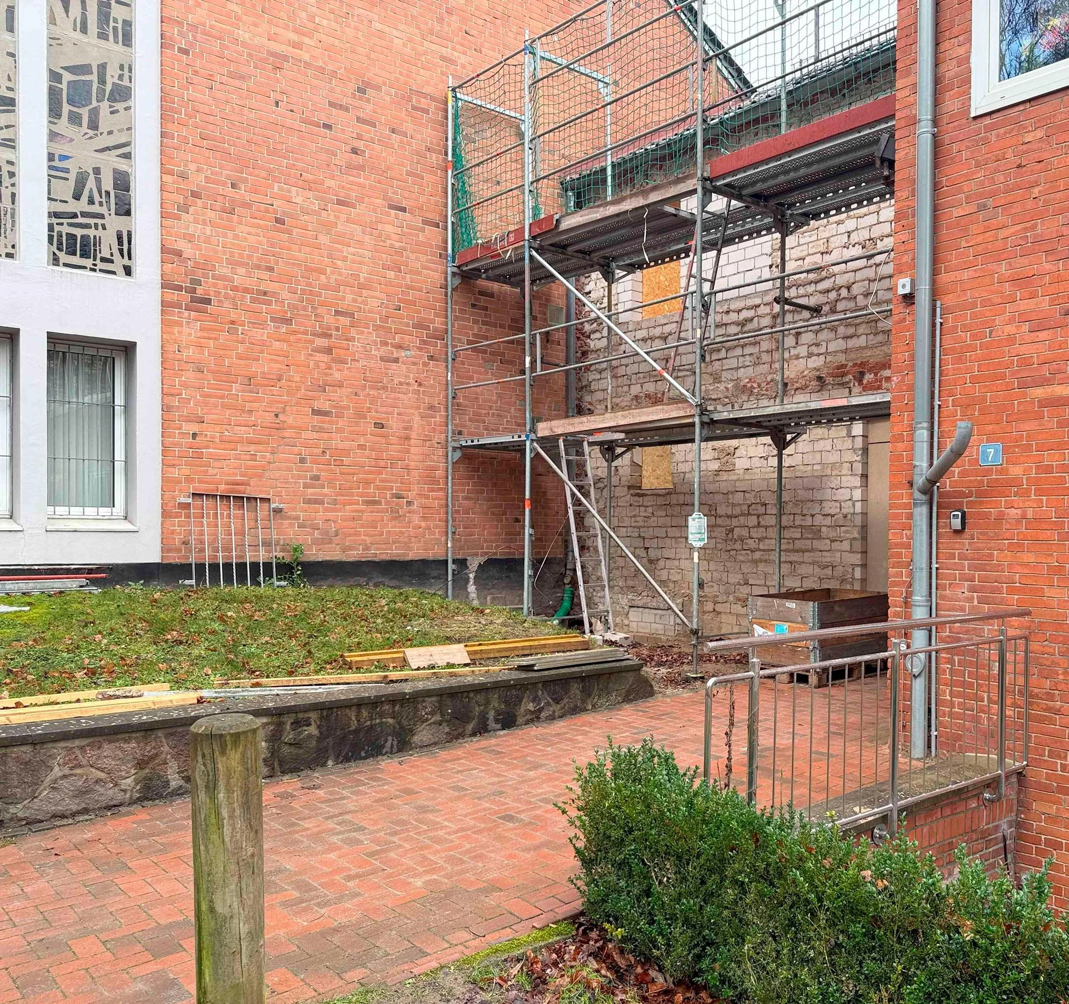 Baustelle an einer roten Backsteinwand mit Gerüst, Bauholz und Bauarbeiten. Es gibt einen kleinen Gartenbereich mit Blumen und eine gepflasterte Fläche.