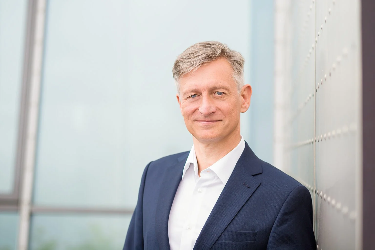 Porträt eines Mannes in einem dunklen Anzug, der an einer Wand mit Schließfächern lehnt.