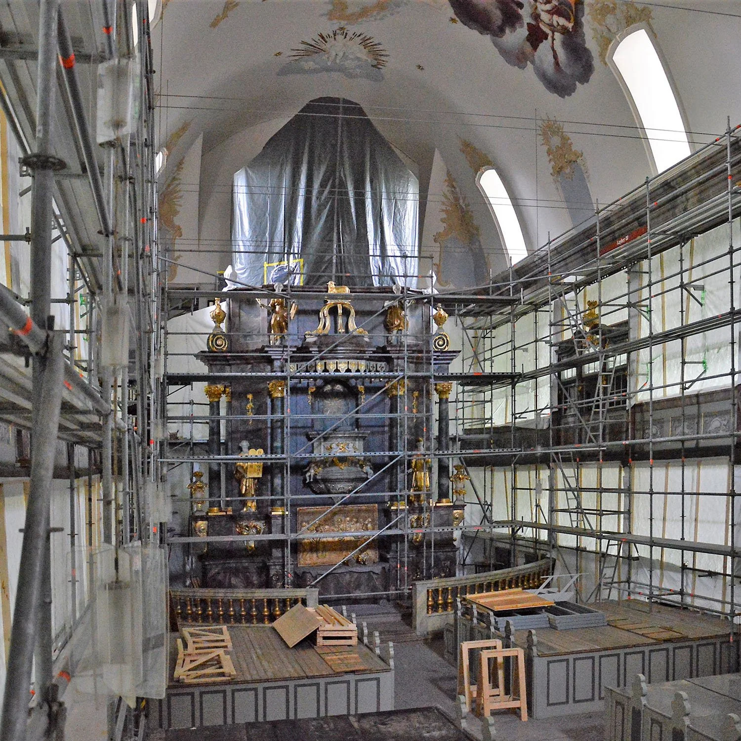 Innenansicht einer Kirche im Renovierungsprozess, mit Gerüsten und Abdeckplanen, im Hintergrund ein Altar mit goldverzierten Statuen und Gemälden, sowie hohen Fenstern und einer bemalten Decke.