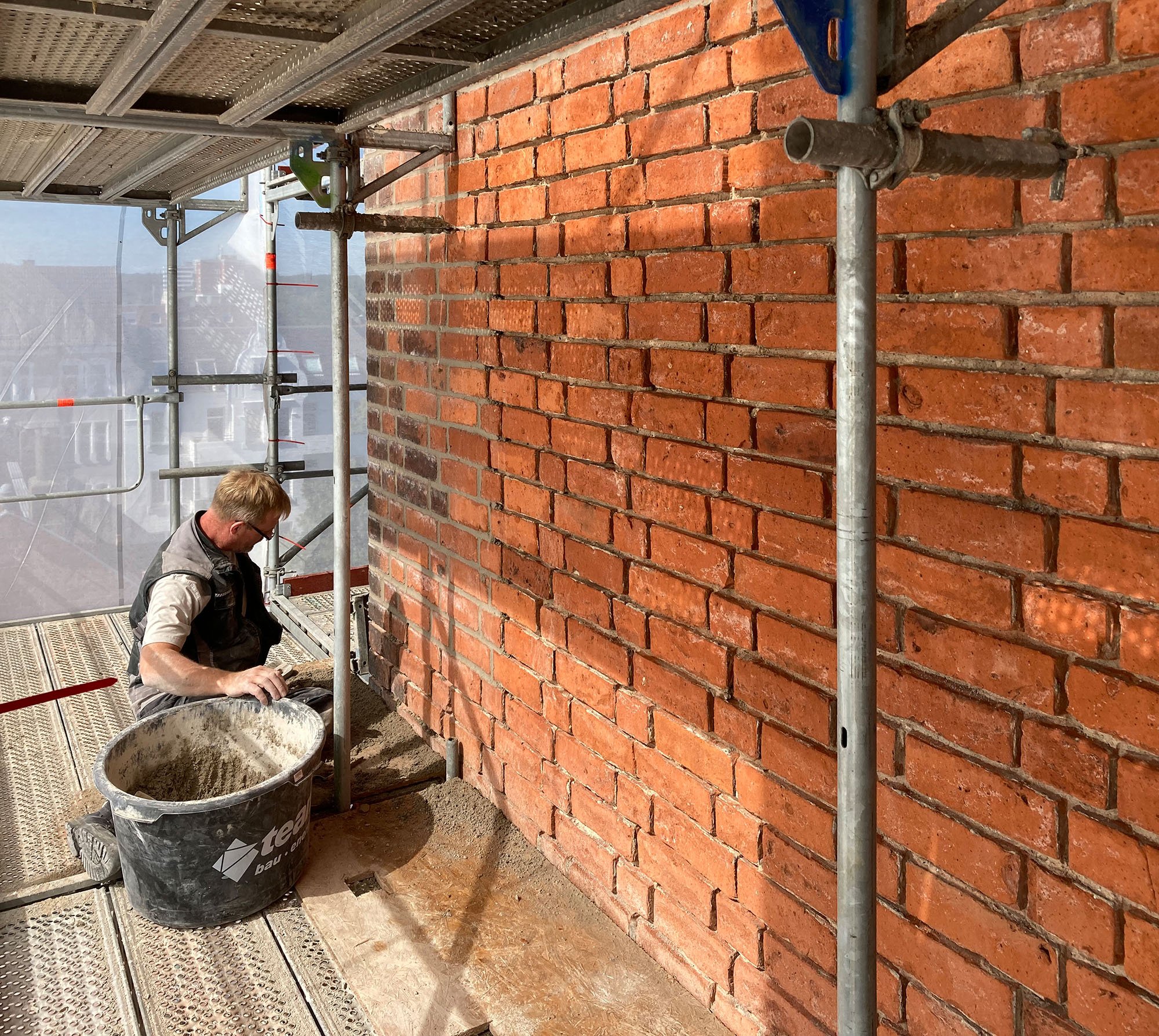 Ein Handwerker arbeitet auf einer Gerüstplattform, während er an einer roten Backsteinmauer arbeitet, mit einem großen Eimer neben sich.