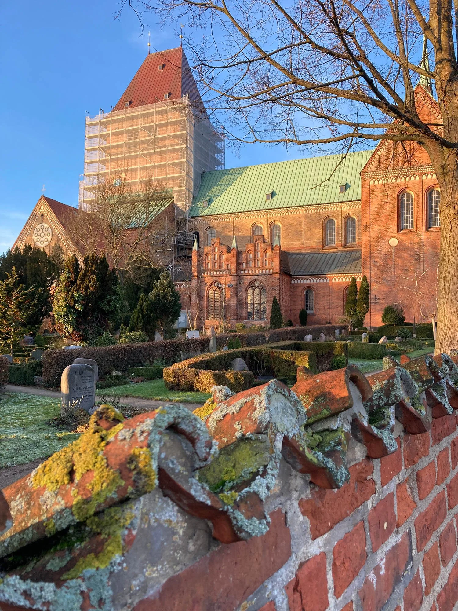 Eine gotische Kirche mit rotem Backstein, teilweise eingerüstet, umgeben von einem Garten mit Zäunen, Steinen und Bäumen bei Sonnenlicht, im Hintergrund blauer Himmel.