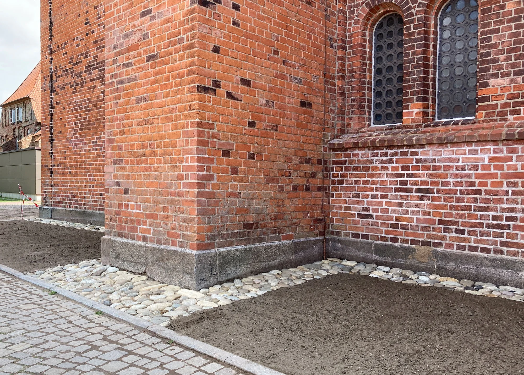 Außenwand eines roten Backsteingebäudes mit zwei hohen, schmalen Fenstern. Der Bereich vor der Wand ist teilweise ausgegraben und mit Steinen und Erde bedeckt.