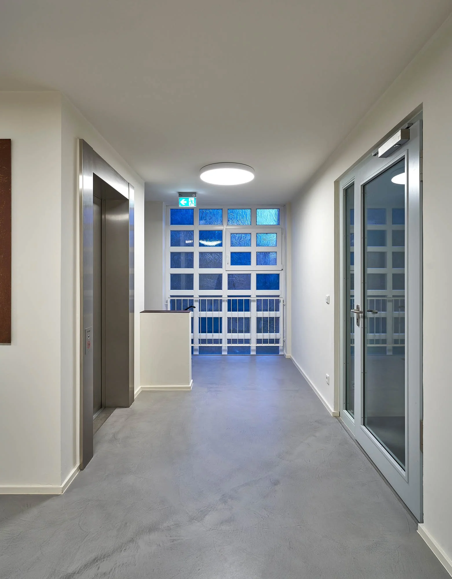 Innenansicht eines modernen Gebäudeflurs mit Aufzug, Glasfenster und Doppeltür, bei Dämmerung oder Nacht.