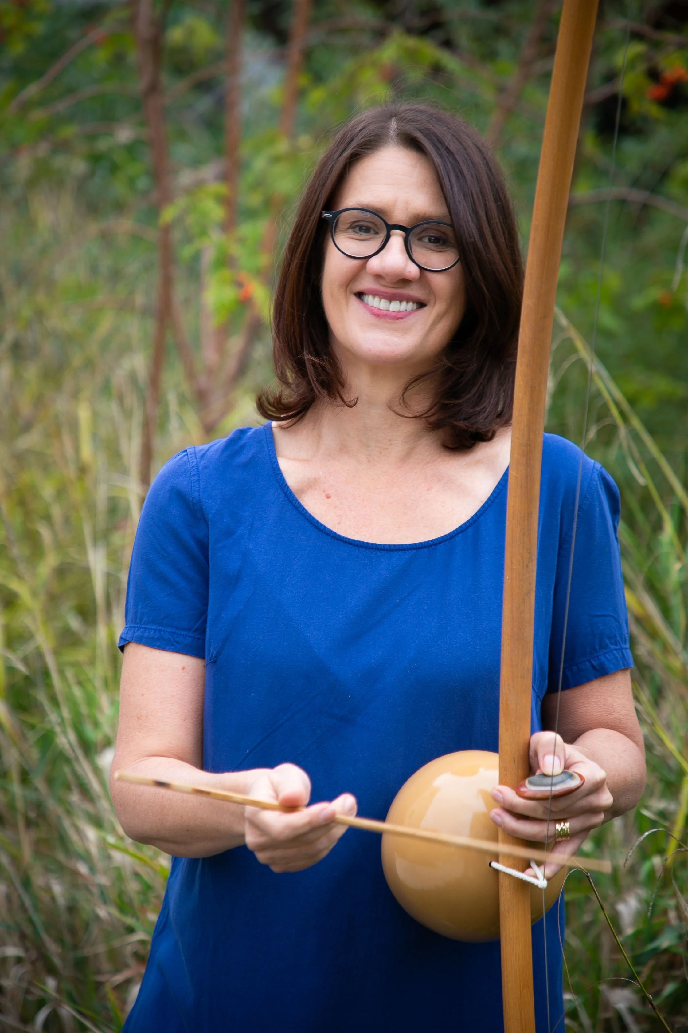Femme souriante tenant un instrument de musique, debout dans un environnement naturel avec des plantes et des arbres en arrière-plan.