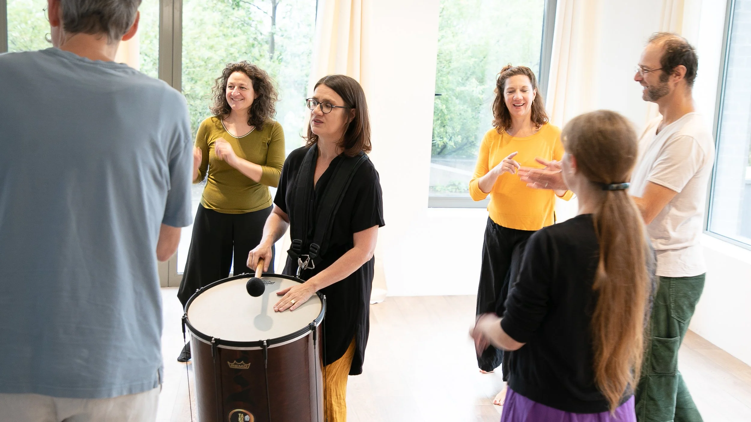 Groupe de personnes joyeuses dans une salle lumineuse, échangeant et regardant une femme avec un tambour.