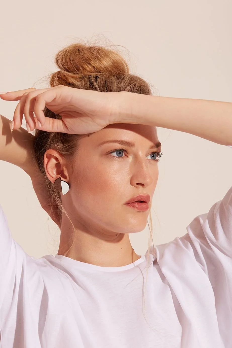 A woman with red hair styled in a bun, wearing a white top and earrings, poses with her arms raised and her hands on her forehead, looking posed against a plain background.