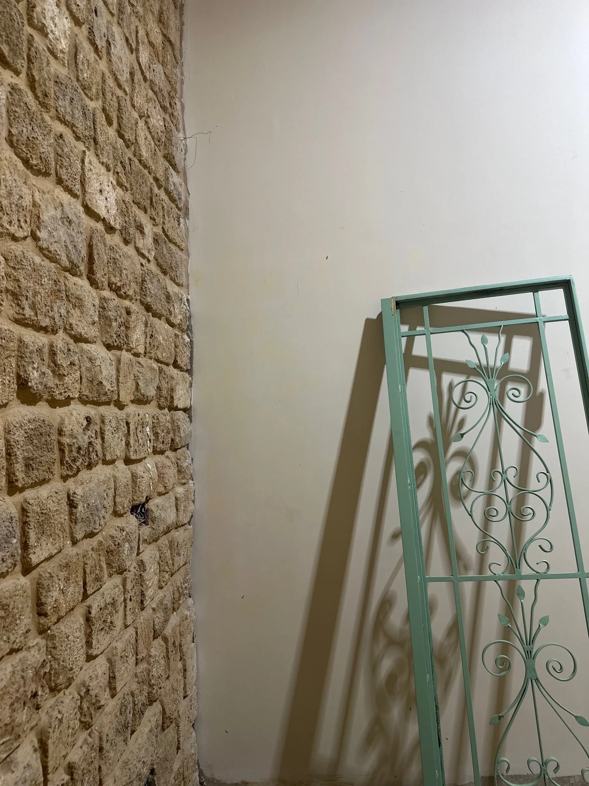 A corner of a room with a exposed brick wall on the left and a plain white wall on the right, with a decorative metal gate leaning against the white wall, casting a shadow.