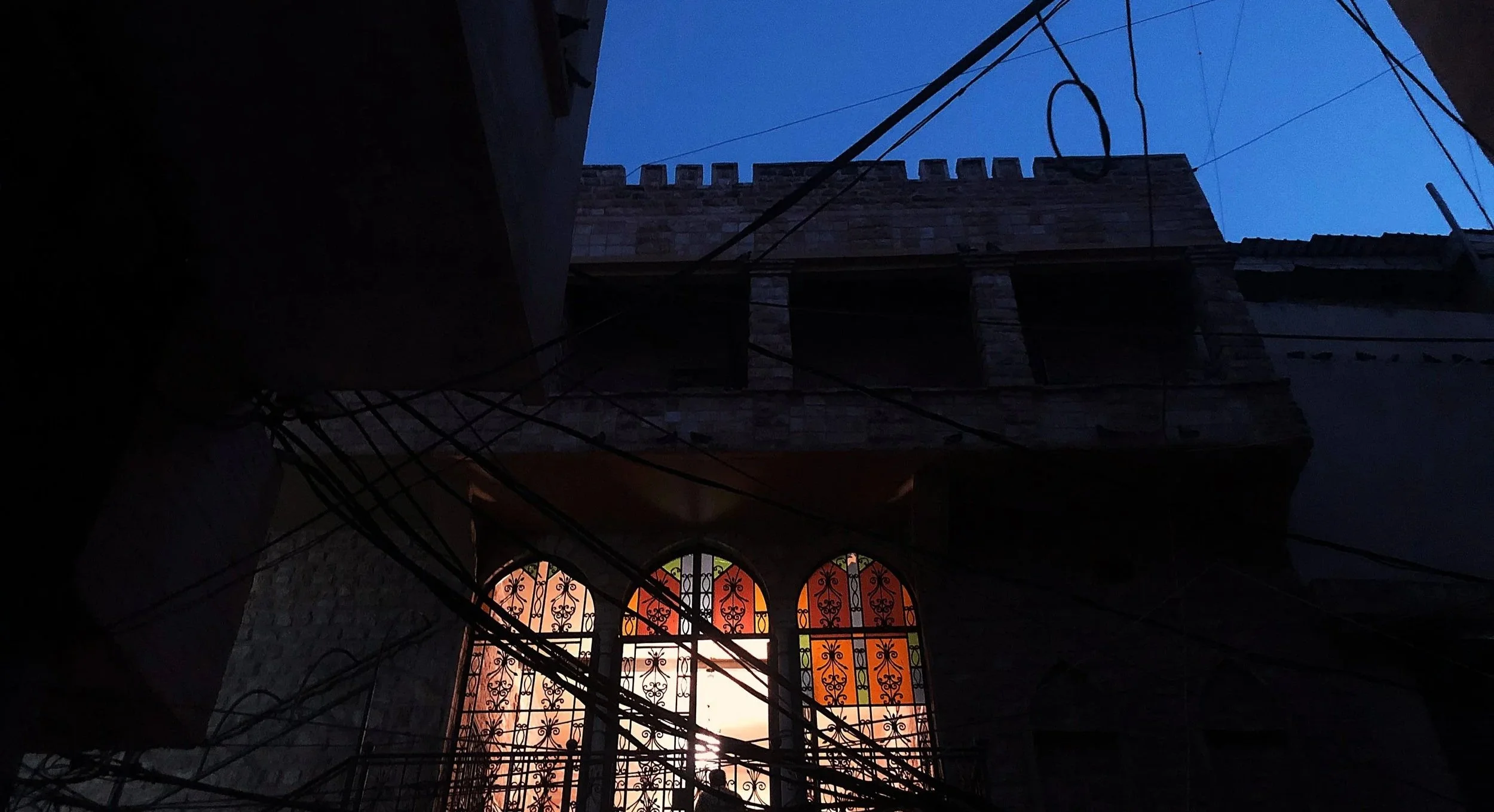 View of a building with stained glass windows and a stone facade, seen from below with wires crisscrossing in front, during dusk or dawn.