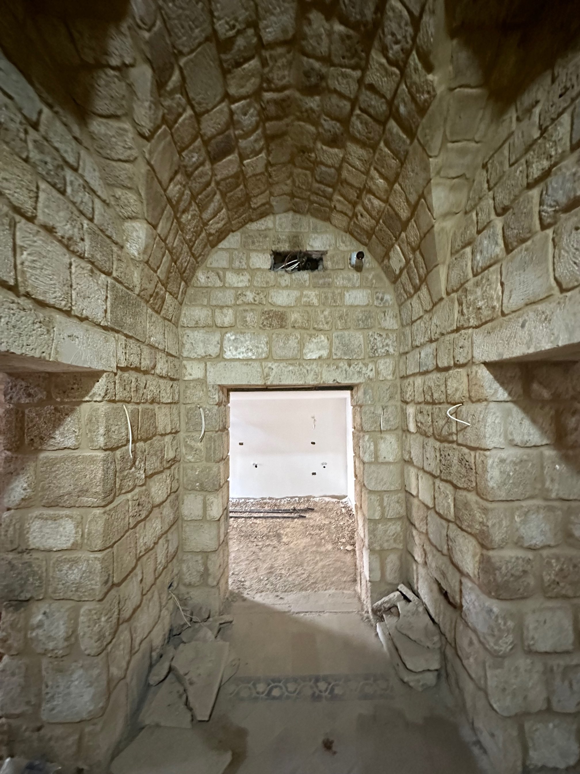 A narrow stone hallway with an arched ceiling under construction. The walls are made of light-colored bricks, and there is a doorway leading to a space with white walls in the background. Construction materials and debris are on the floor.