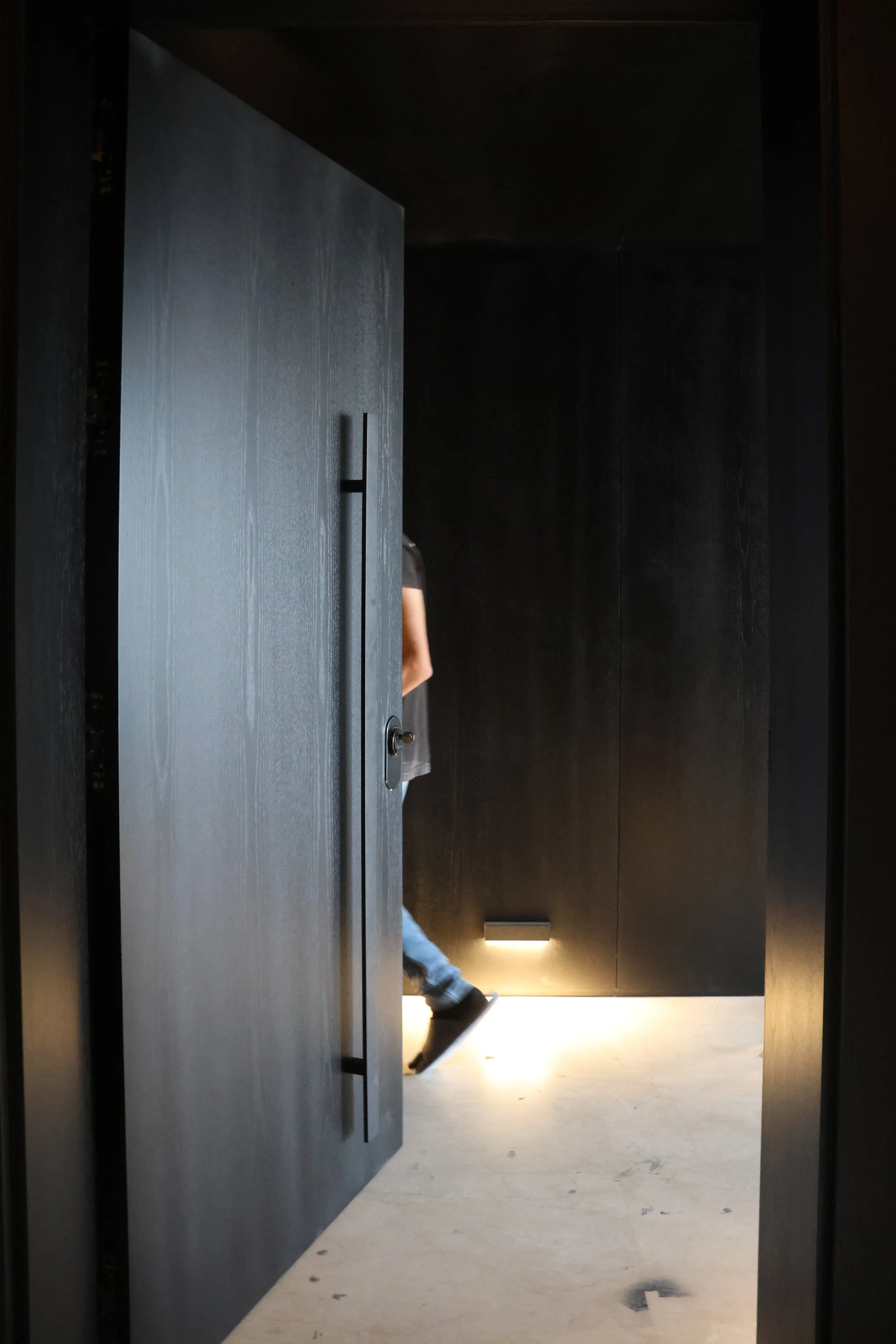 A person entering a dark room through a slightly open wooden door, with only part of their leg and arm visible.