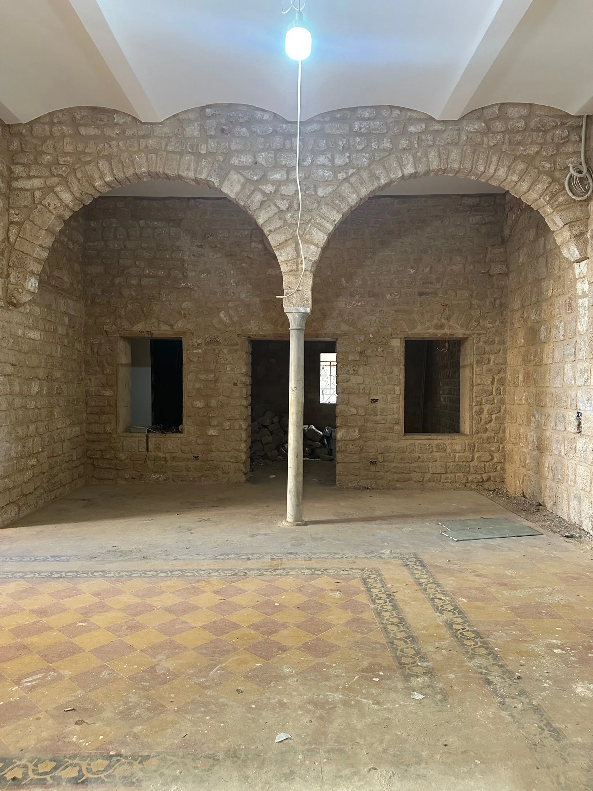Interior of a building under construction with brick arches, stone walls, and a partially finished patterned tile floor. A pole is centered supporting a hanging light fixture.
