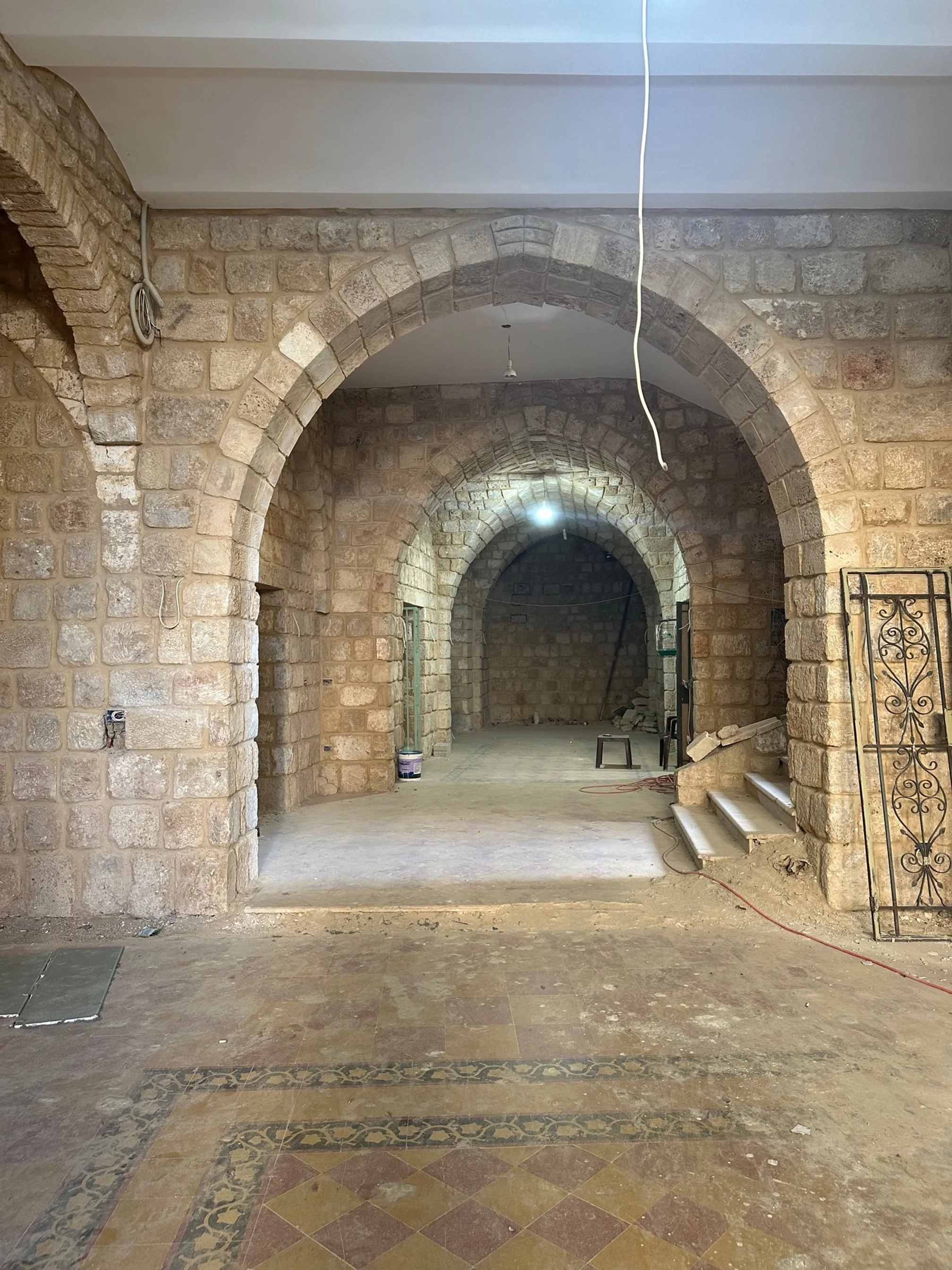 Inside a building with stone archways, unfinished flooring, construction tools, and materials, indicating ongoing renovation.