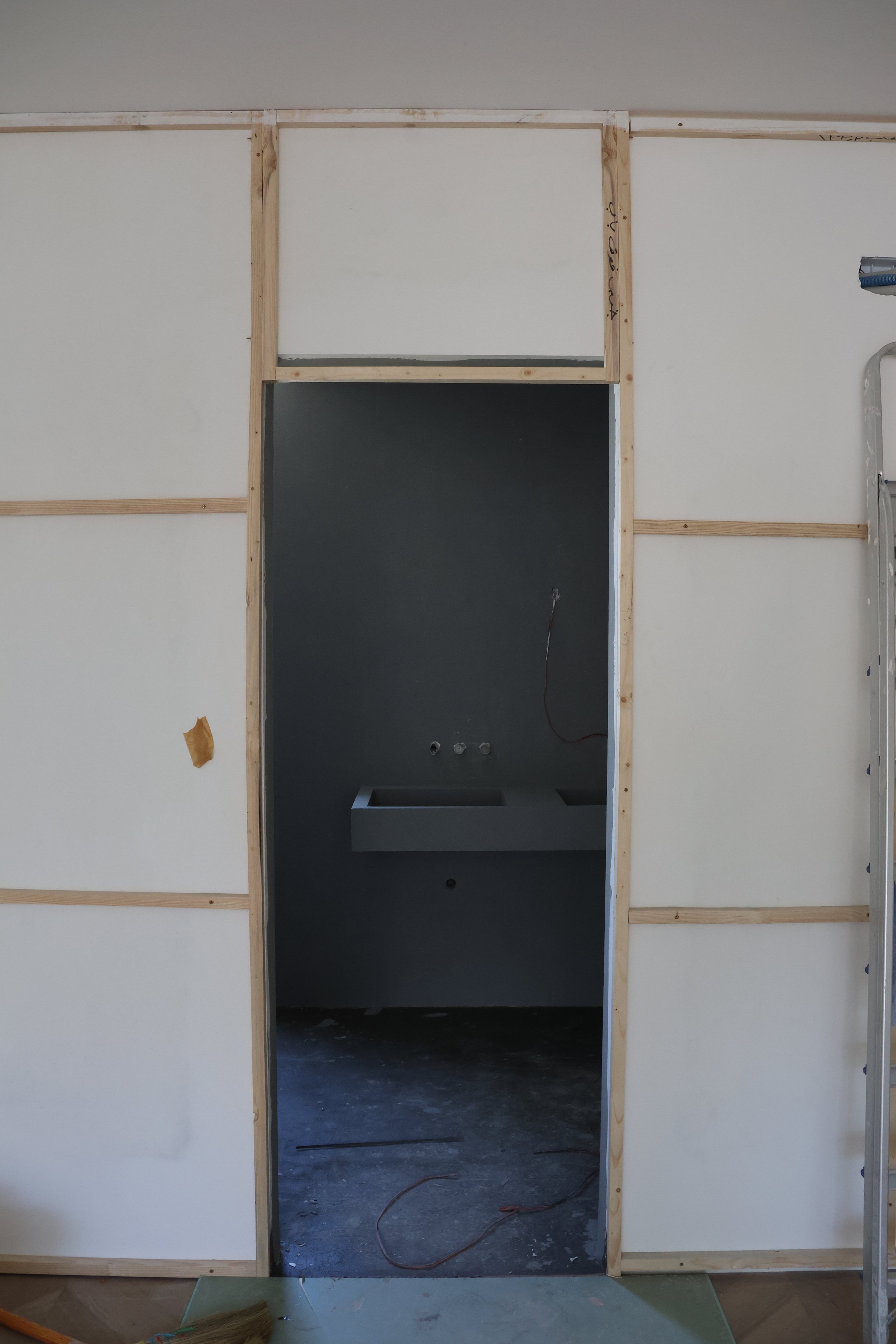 Construction site showing a doorway frame in progress, with exposed wooden studs and a dark room with a sink in the background.