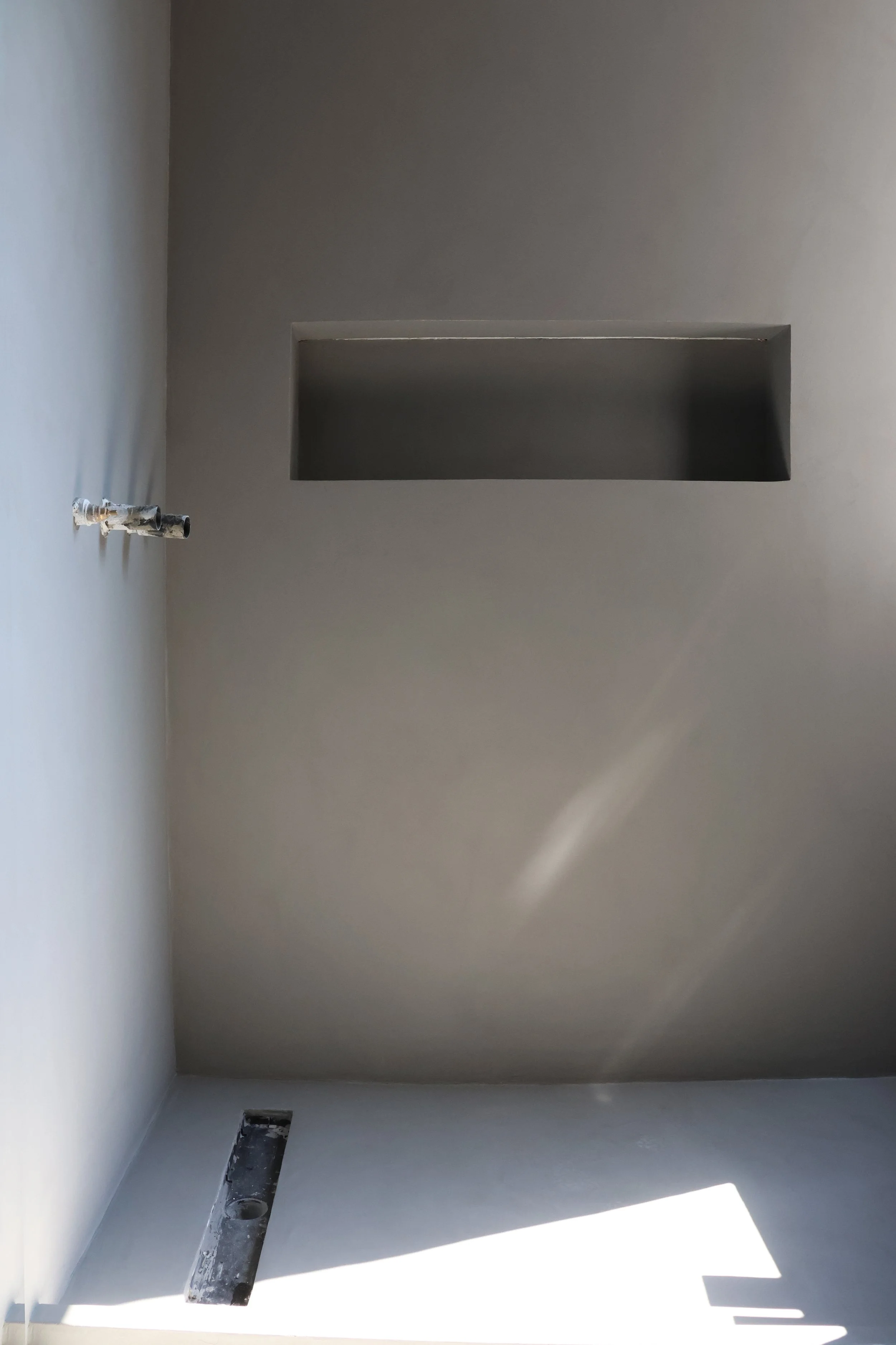 Interior view of a partially finished room with light gray walls, a rectangular recessed shelf, exposed plumbing, and a floor drain, with sunlight casting shadows.