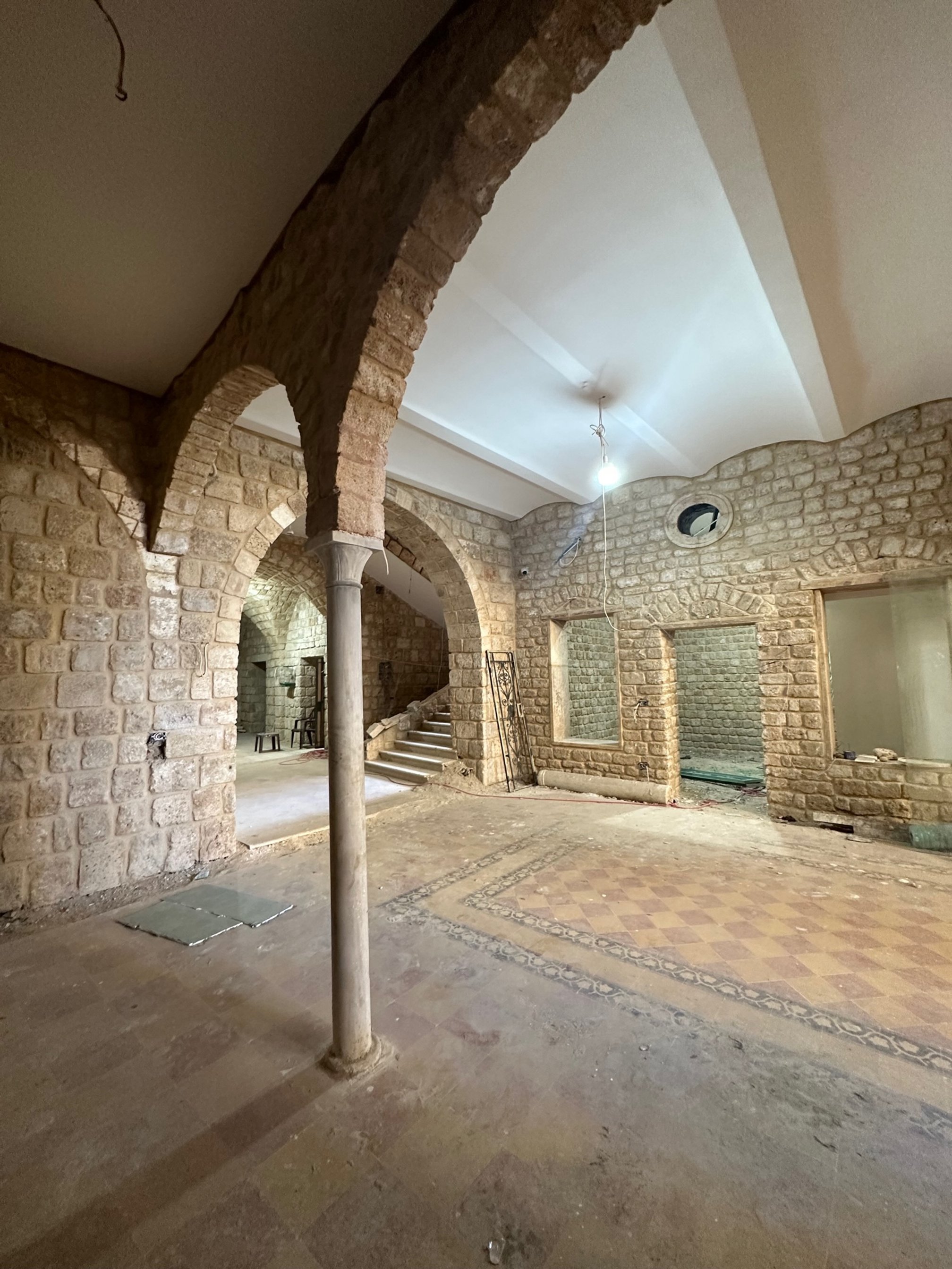 Interior of a building under construction with stone archways, brick walls, and tiled flooring, unfinished lighting, and construction materials scattered.