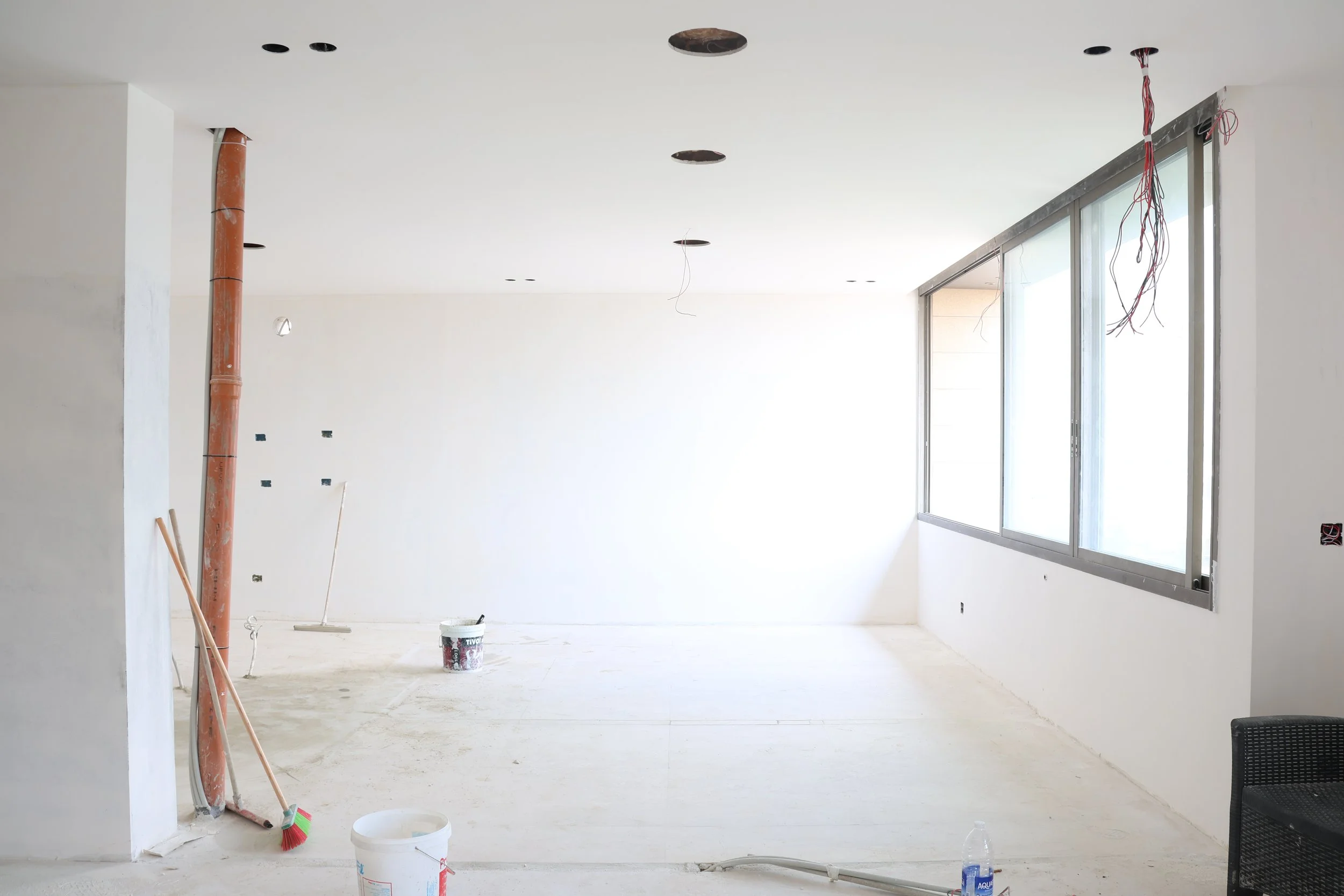 Empty room under construction with unfinished walls, electrical wires hanging from the ceiling, and construction tools on the floor near a large window.