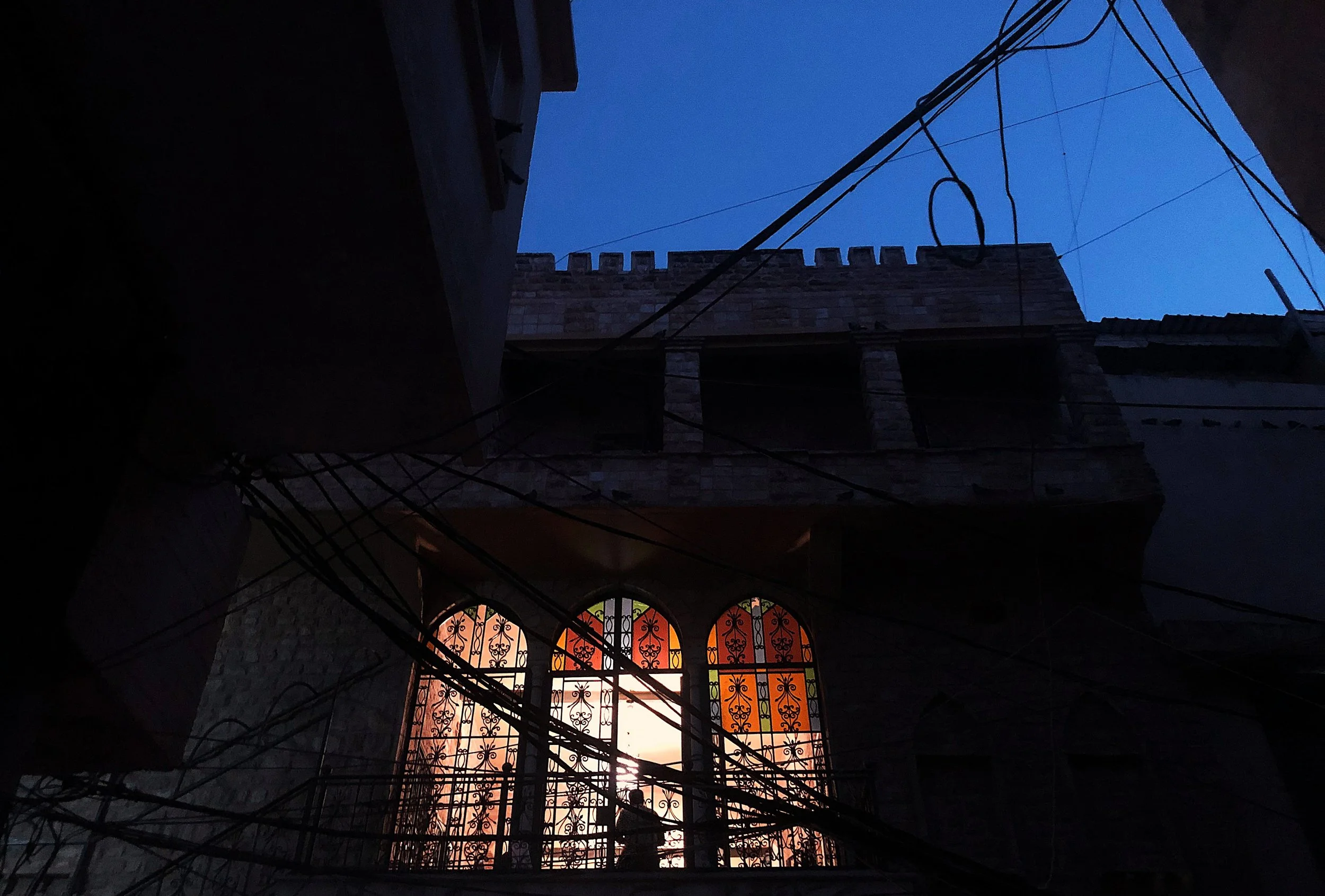 View of the sky at dusk seen through an alleyway with dark silhouetted buildings, electrical wires crossing in front, and a decorative wrought iron window with illuminated glass panes at the bottom.