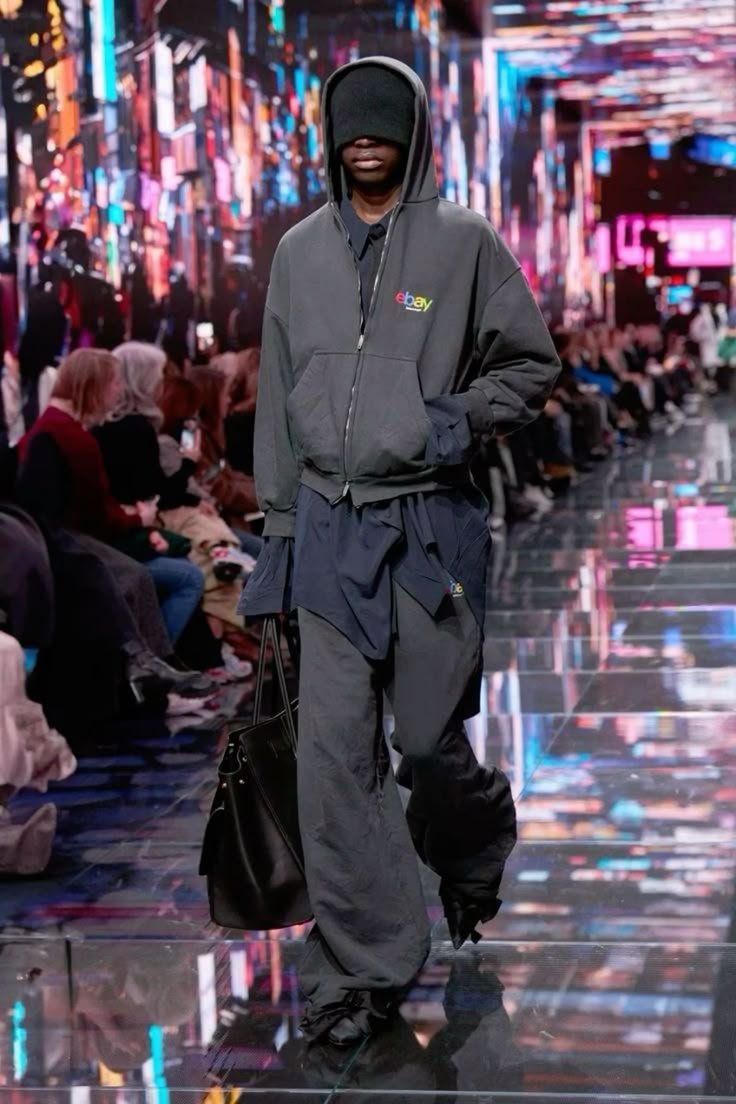 A model walking on a runway wearing a black hooded sweatshirt with the eBay logo, baggy black pants, and holding a black handbag. The model's face is partially covered with the hood, and the background features a colorful, digital-themed backdrop with seated audience members.