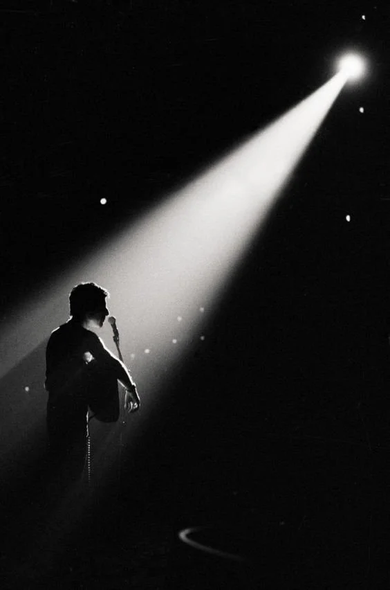 Silhouette of a person singing or speaking into a microphone on stage, illuminated by a bright spotlight against a dark background.