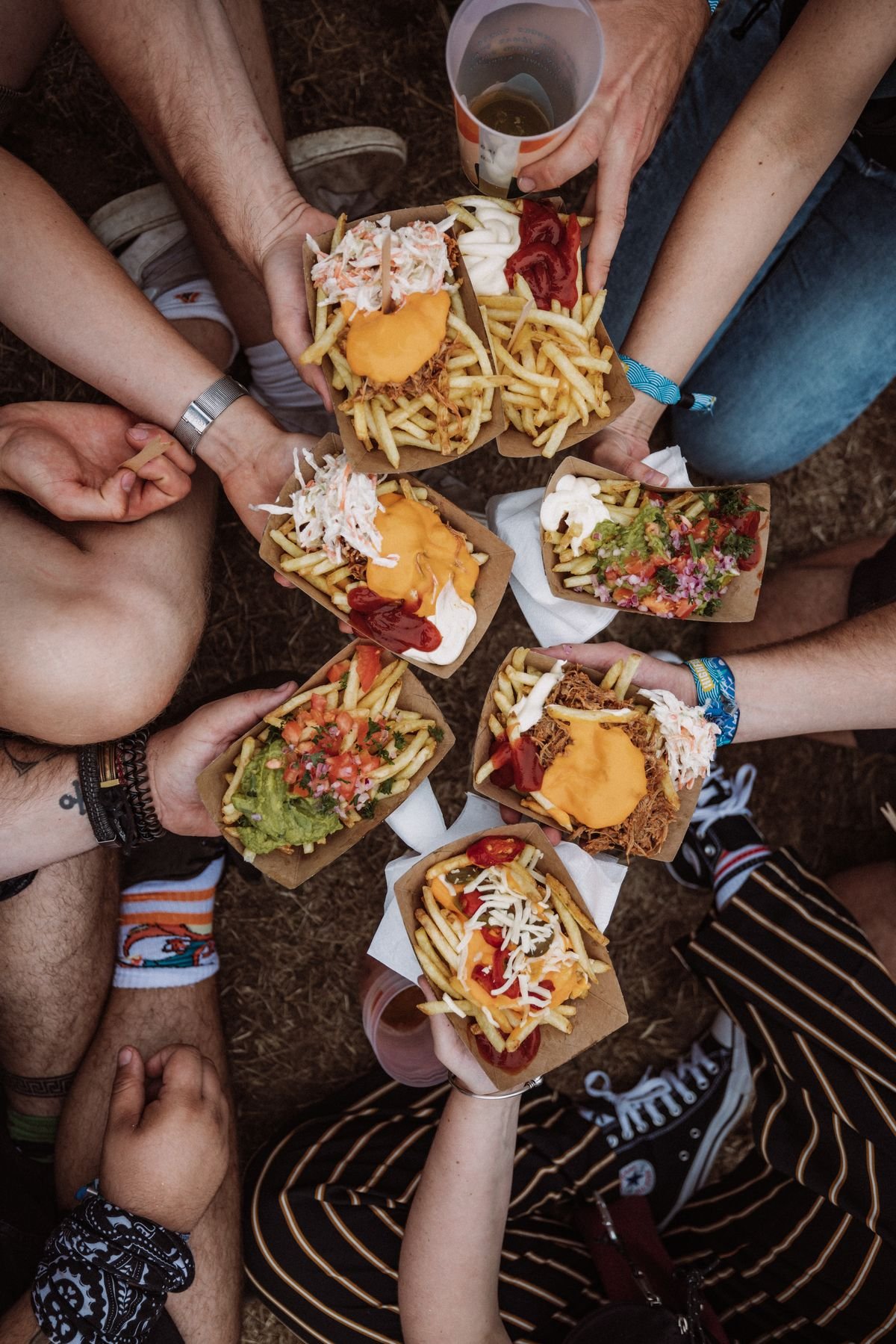 Group of people sitting on the ground holding paper trays of loaded French fries with cheese, sour cream, shredded cheese, salsa, shredded chicken, and toppings, with drink cups visible.
