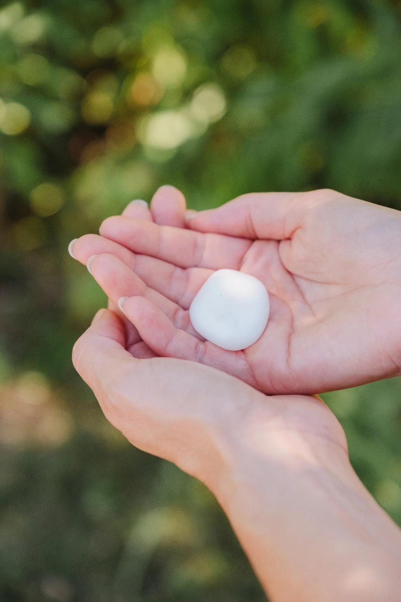 Person holding a white, round stone in their hand outdoors with green foliage in the background.