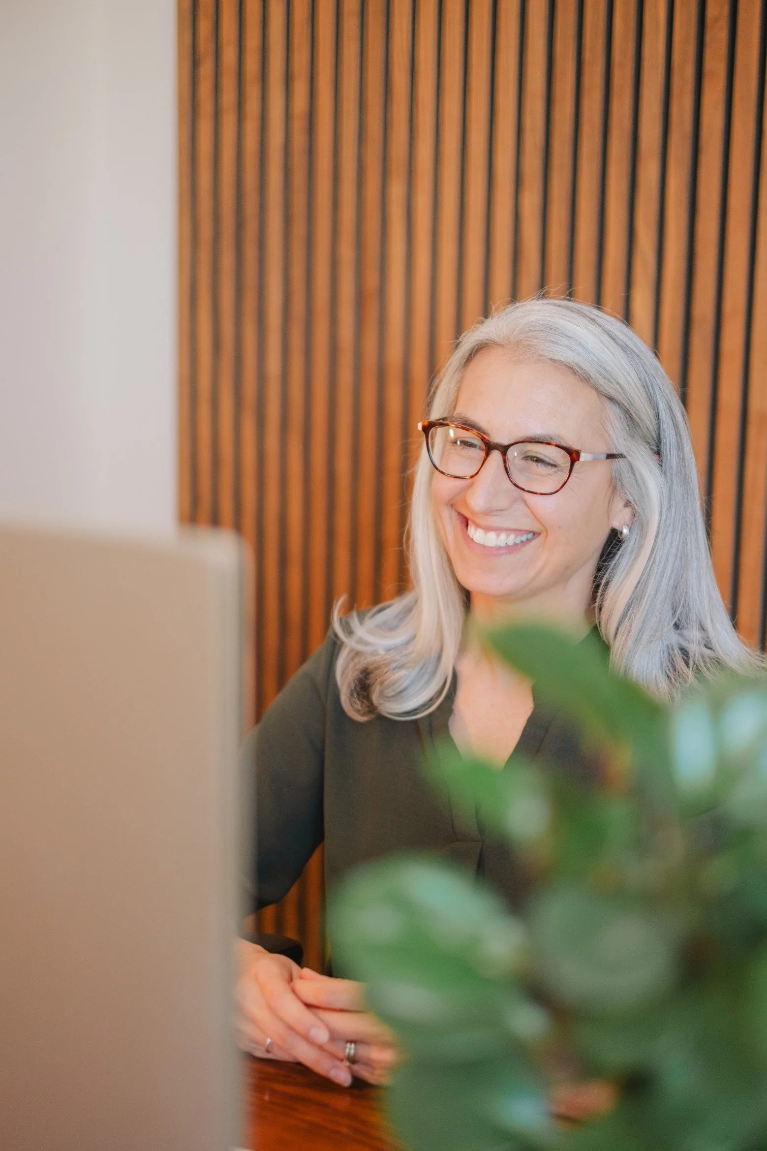 A smiling woman with gray hair and glasses sitting at a table in front of a laptop, with a plant partially visible in the foreground and a wooden wall in the background.