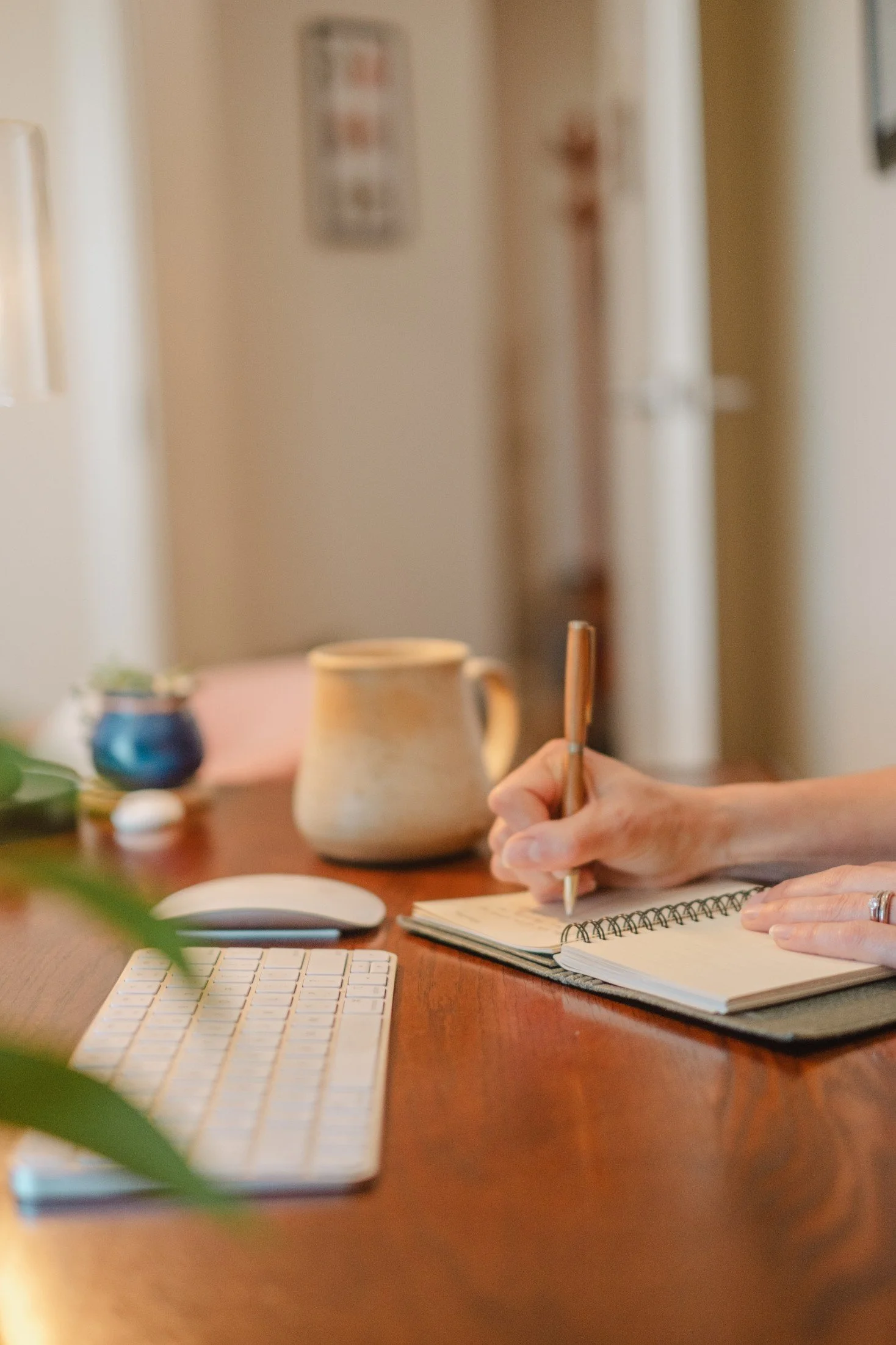 A person writing in a spiral notebook at a wooden desk with a computer keyboard and mouse, a mug, and decorative plants and objects in the background.