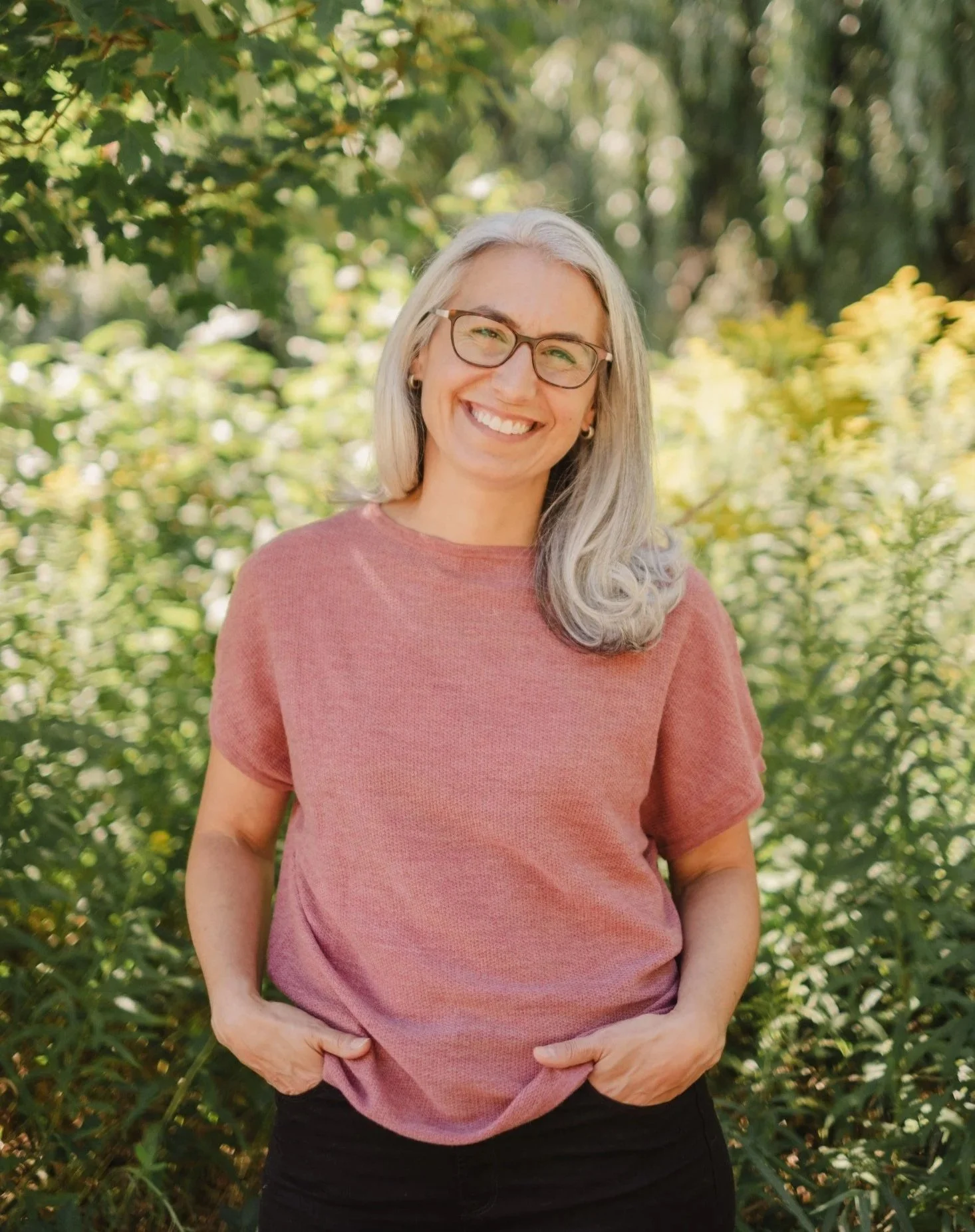 A smiling woman with gray hair, glasses, and earrings standing outdoors surrounded by green foliage.