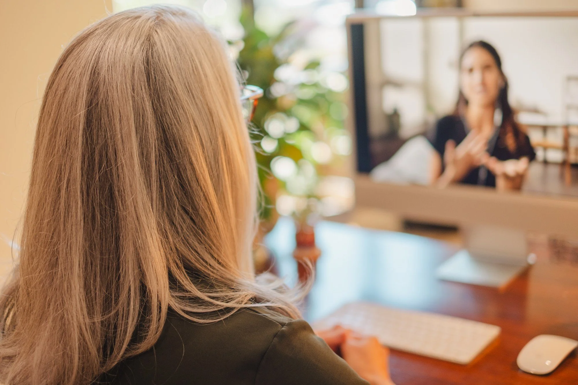 A woman with blonde hair wearing glasses and a dark top is sitting at a desk, engaging in a video call with another woman who appears on a computer screen in the background.