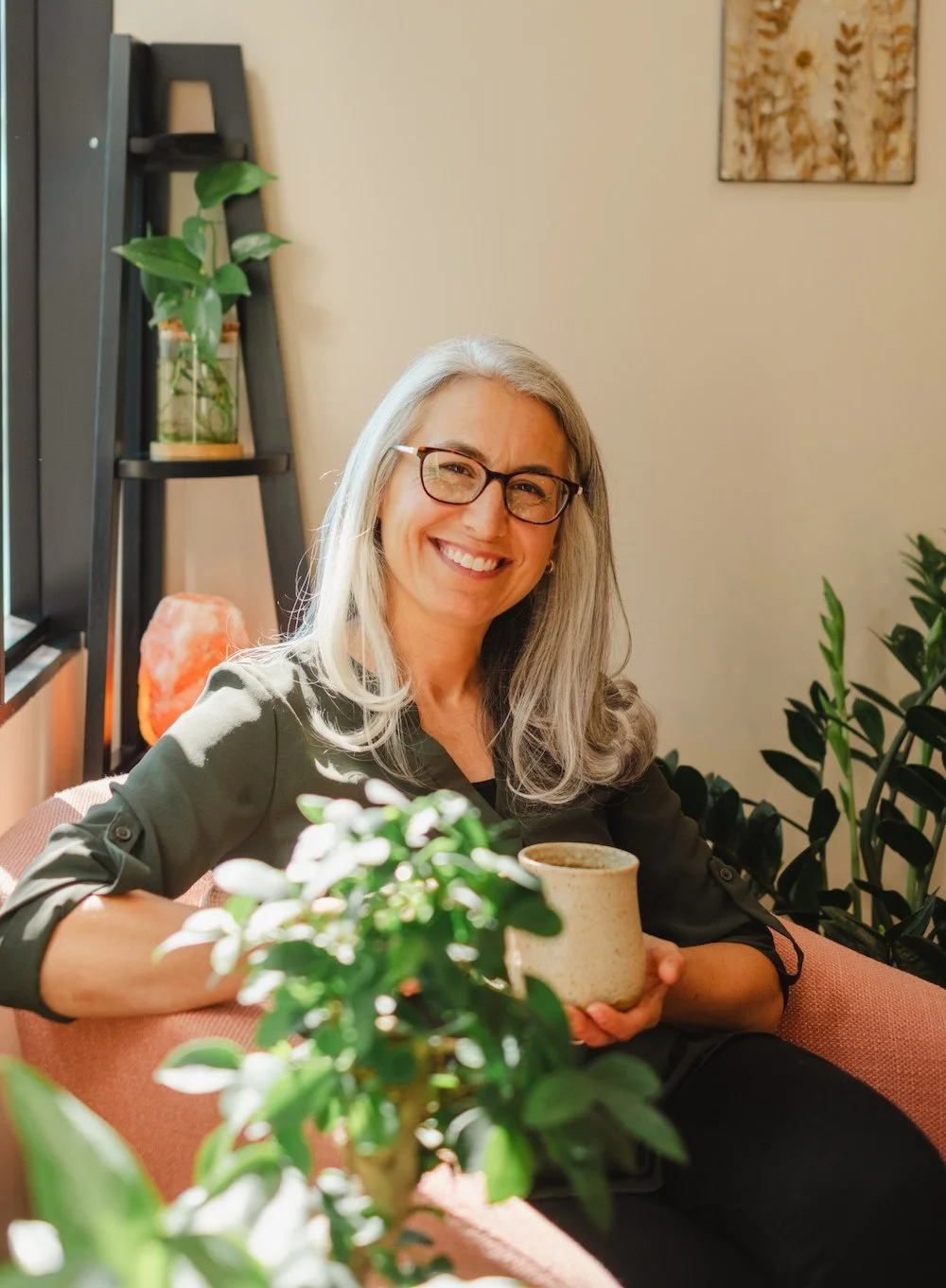 A smiling woman with gray hair and glasses sitting on a pink sofa, holding a cup, surrounded by plants in a cozy, well-lit room.