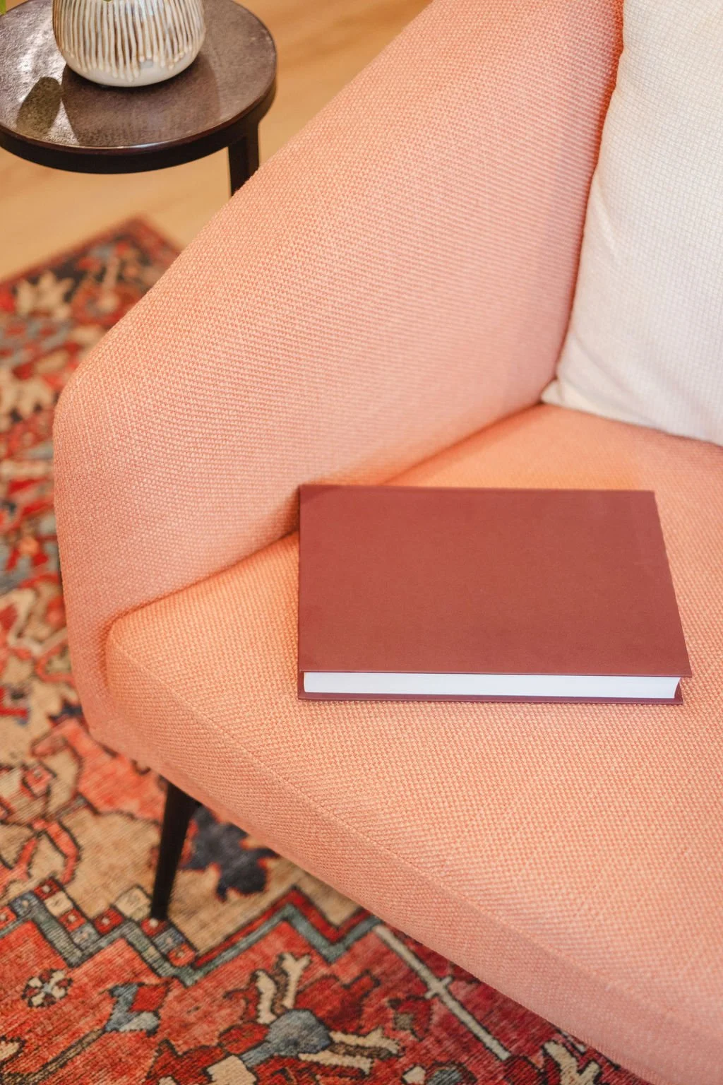 A closed red-brown book resting on a pink upholstered armchair with a white pillow behind it. In the background, there is a round wooden side table with a decorative white and gold ceramic vase on top, and a patterned rug underneath.