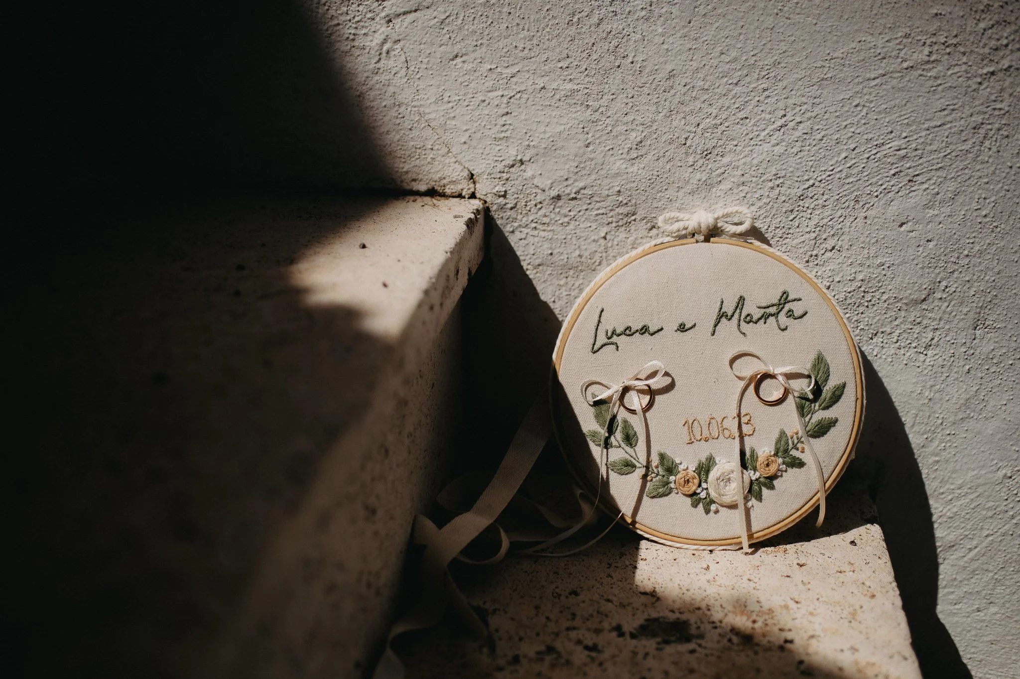 Embroidery hoop with wedding rings, embroidered flowers, and the text "Luca e Marta" and the date "10.06.13", resting on a stone ledge against a white textured wall.