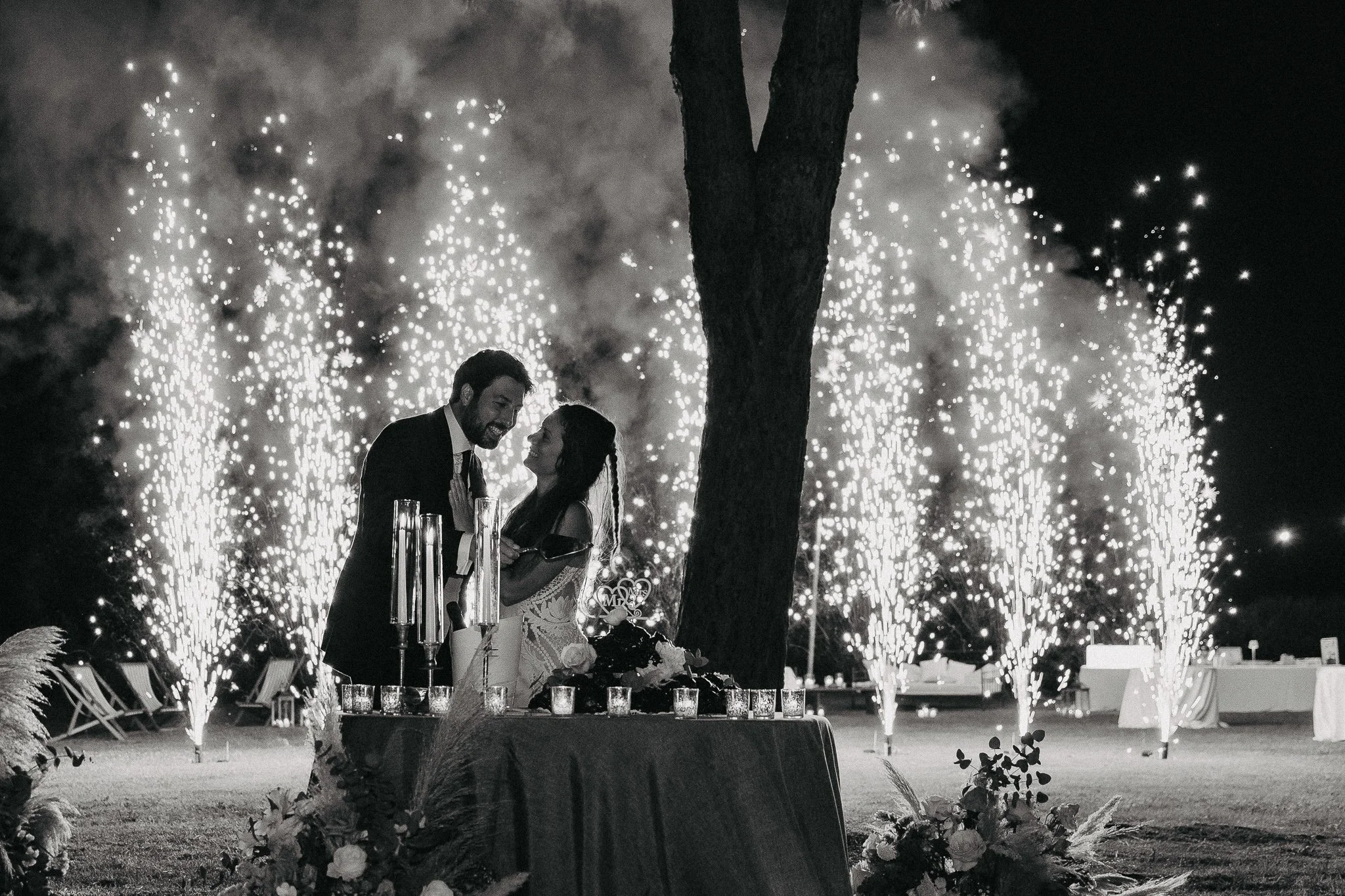 A couple at a wedding reception celebrating with fireworks in the background at night, with a decorated table in front.