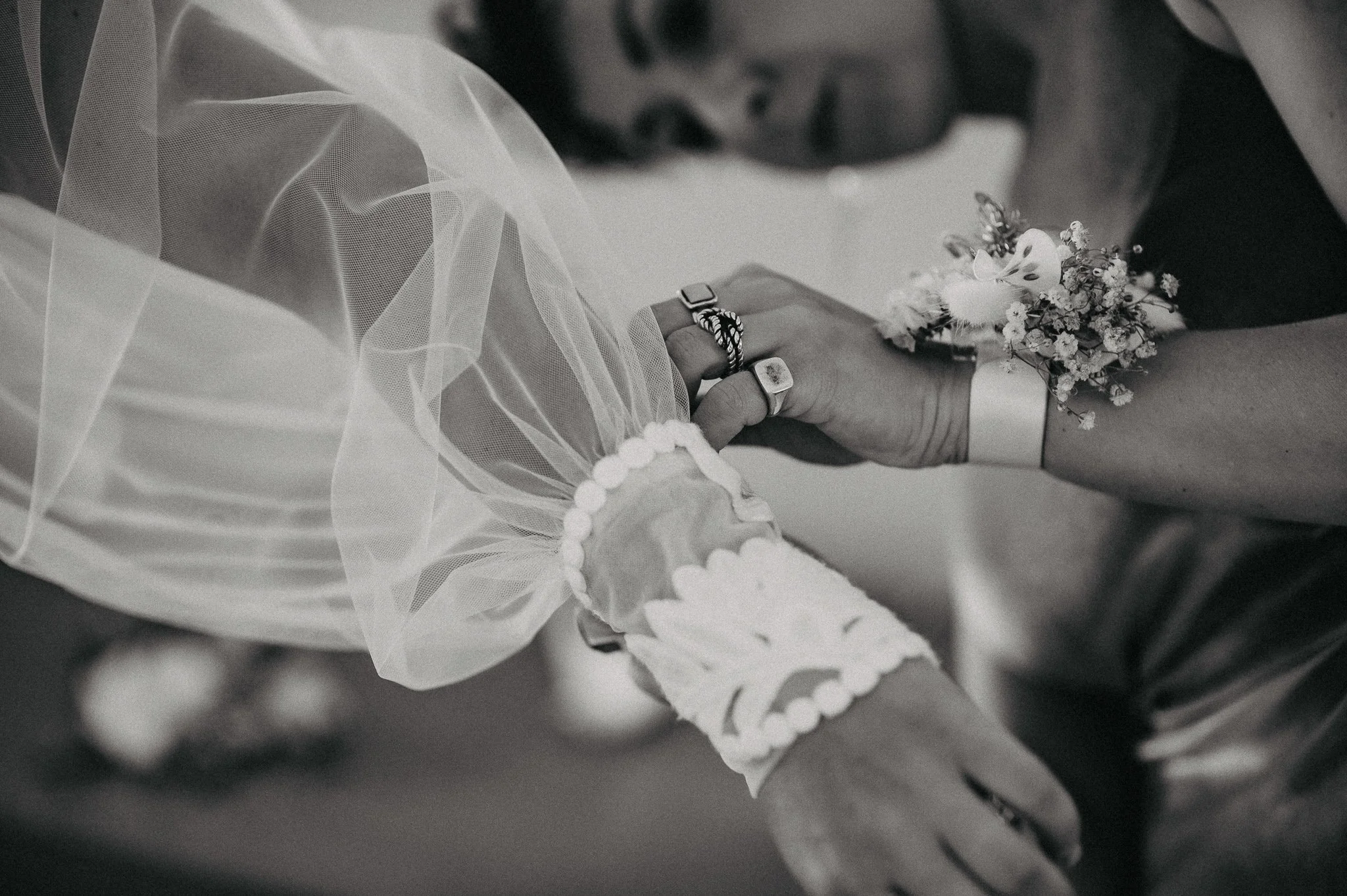 Close-up of two people holding hands during a wedding, wearing rings, with one person wearing a lace and satin sleeve and a floral wrist corsage.