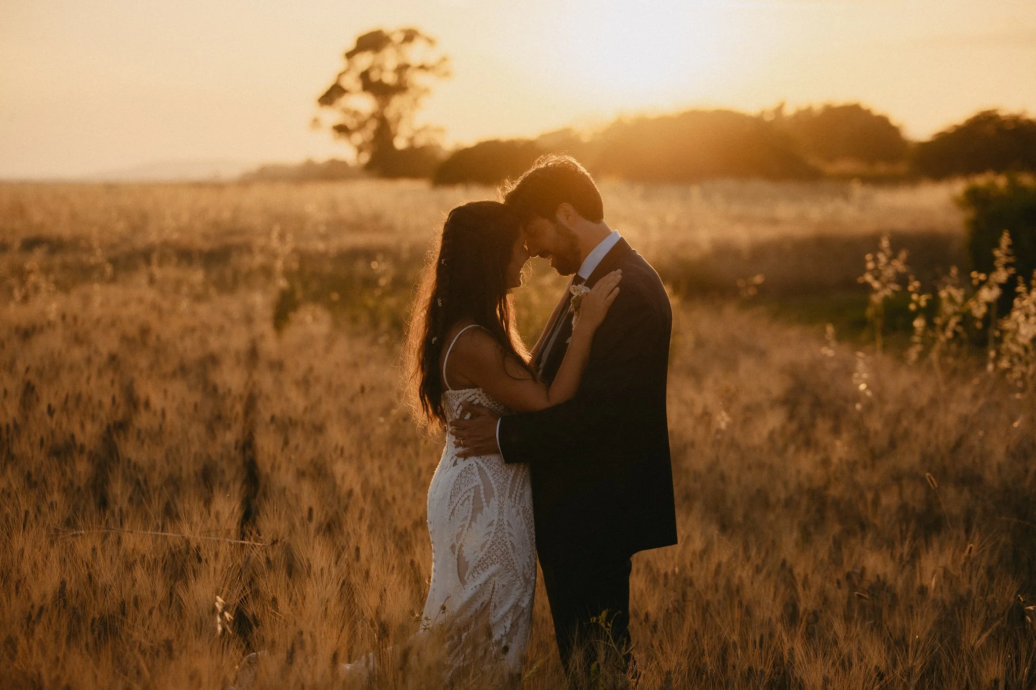 A couple in wedding attire embracing in a field during sunset.