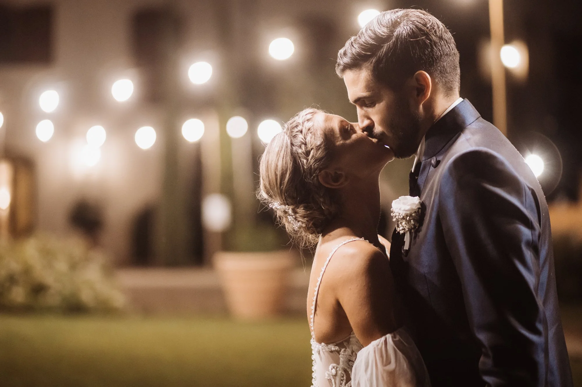 A bride and groom share a kiss at night, dressed in wedding attire under soft lights.