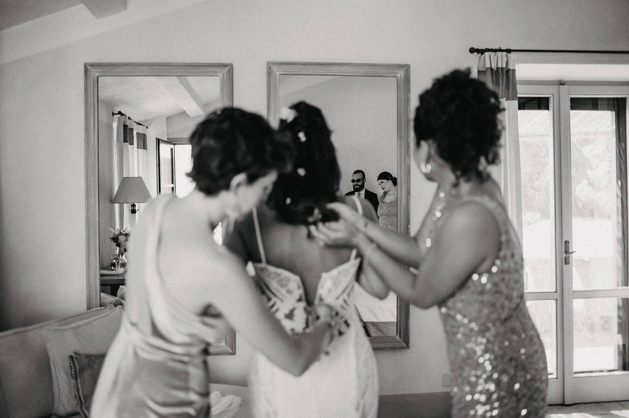 Three women, one in a wedding dress, helping the bride get ready, with a mirror reflecting a man and woman in the background