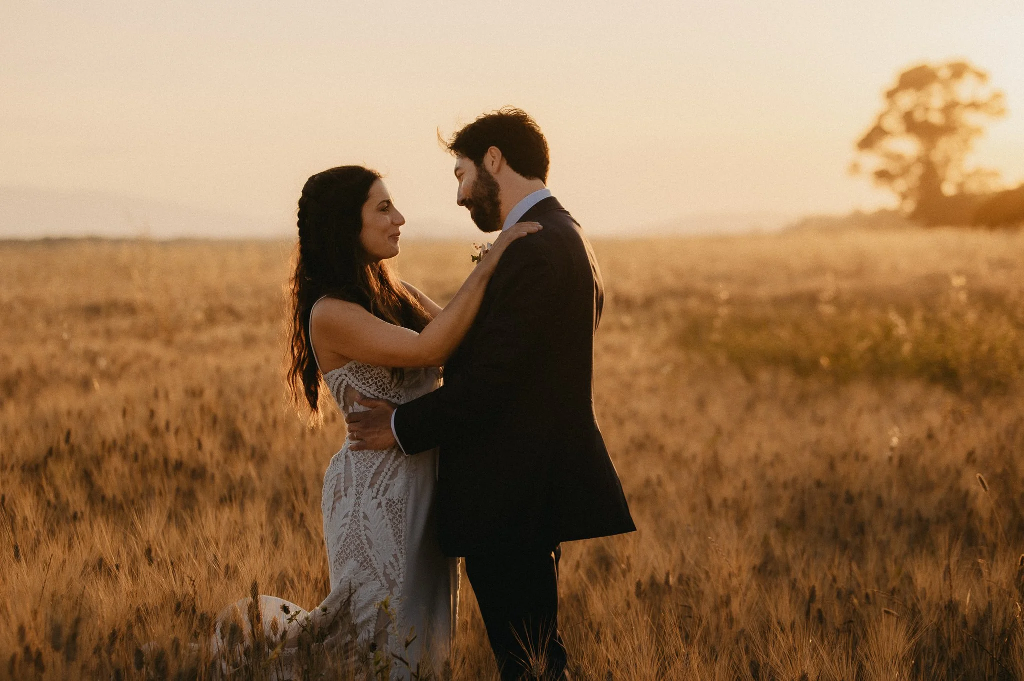A couple in wedding attire embracing in a field at sunset.